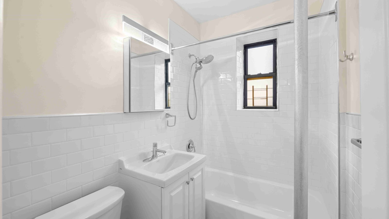 Photograph of a small bathroom with white subway tile, a white vanity sink, a mirror with a light bar above it, a handheld shower head and a bathtub under a small window
