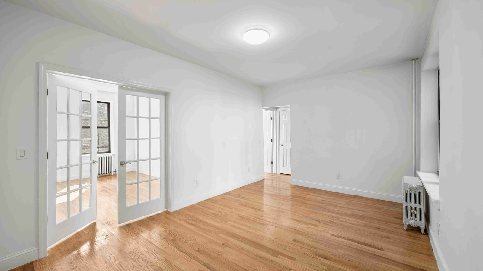 Photograph of an empty, freshly painted white room with a light wood floor, a round ceiling light, a radiator under a window on the right, a doorway to a hall at the back, and a pair of glass-paned French doors open on the left to a second room with a window and another radiator