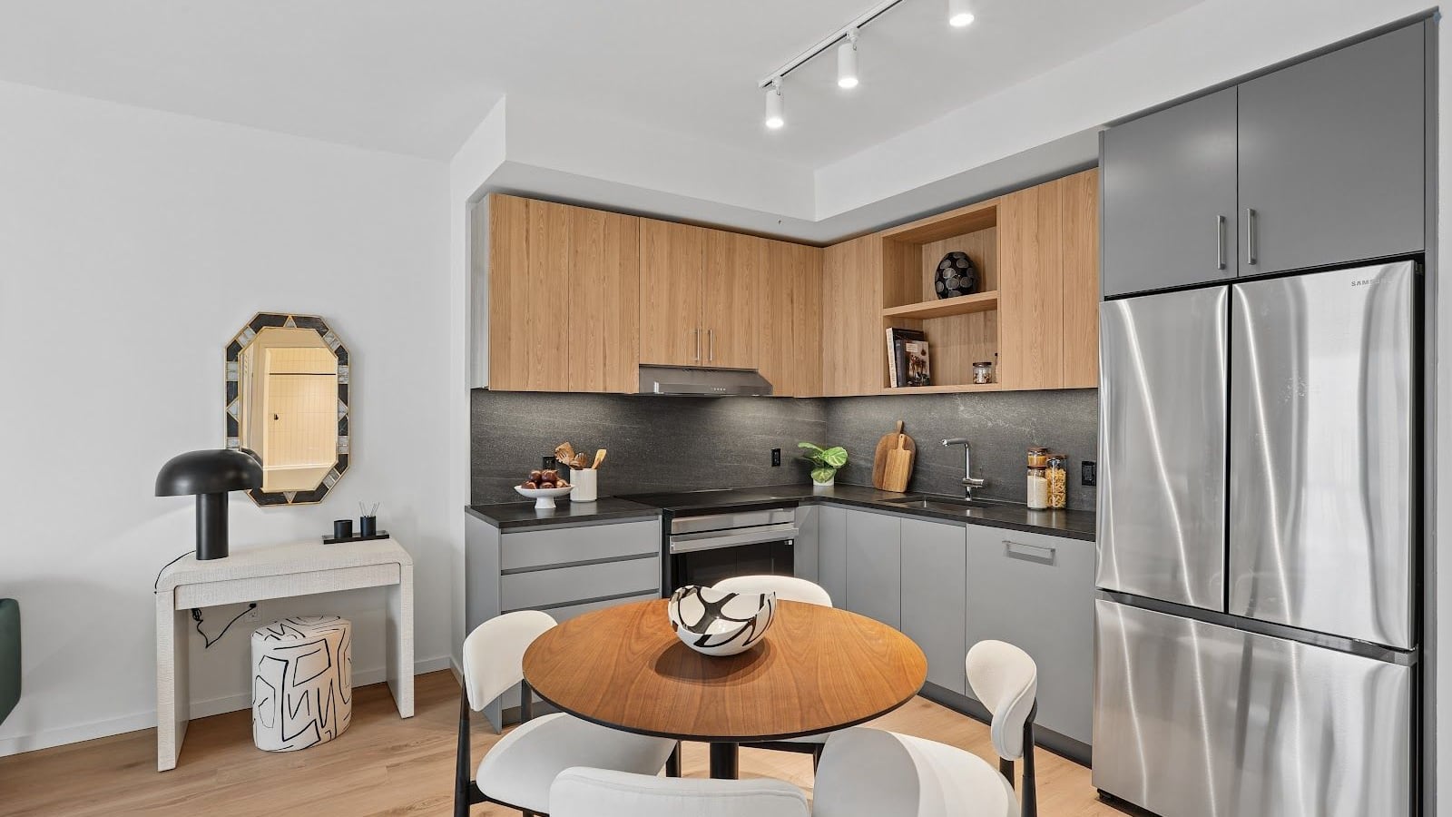 Photograph of an L-shaped kitchen with wood-tone upper cabinets, grey lower cabinets, a dark stone-look counter and backsplash, a smooth-top range with a hood above it, a sink, a stainless-steel French door refrigerator, track lights on the ceiling, and a round wooden dining table with four white chairs in front