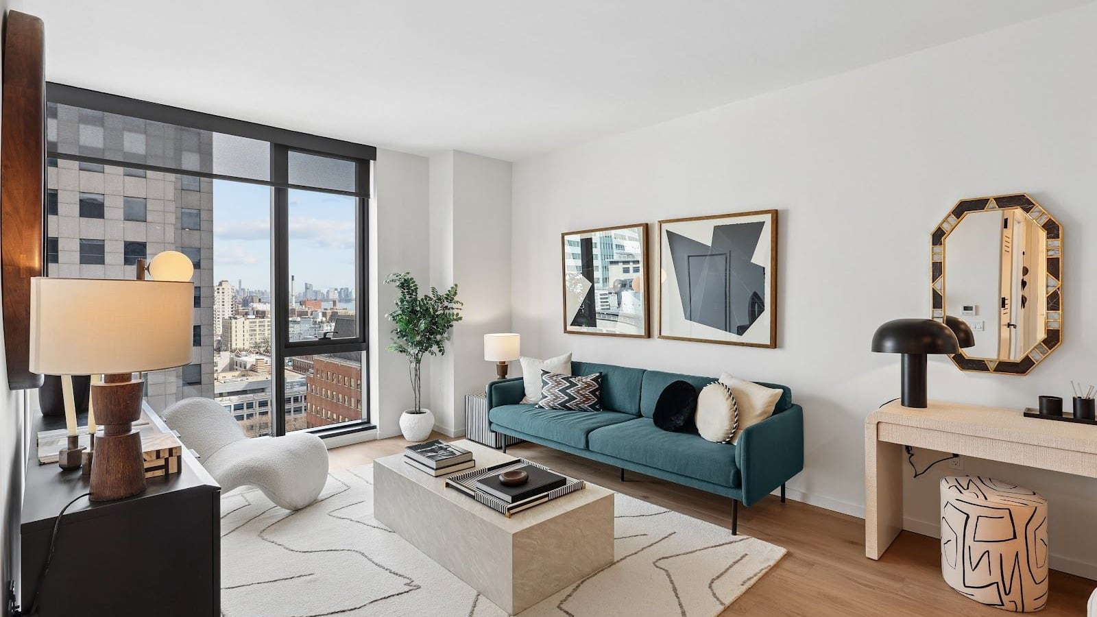 Photograph of a furnished living room with pale wood flooring and white walls, a teal sofa, a stone-colored coffee table on a patterned rug, a white lounge chair, two framed prints, an octagonal mirror above a console table, and a floor-to-ceiling window with a dark frame looking past an office building to rooftops and a distant skyline