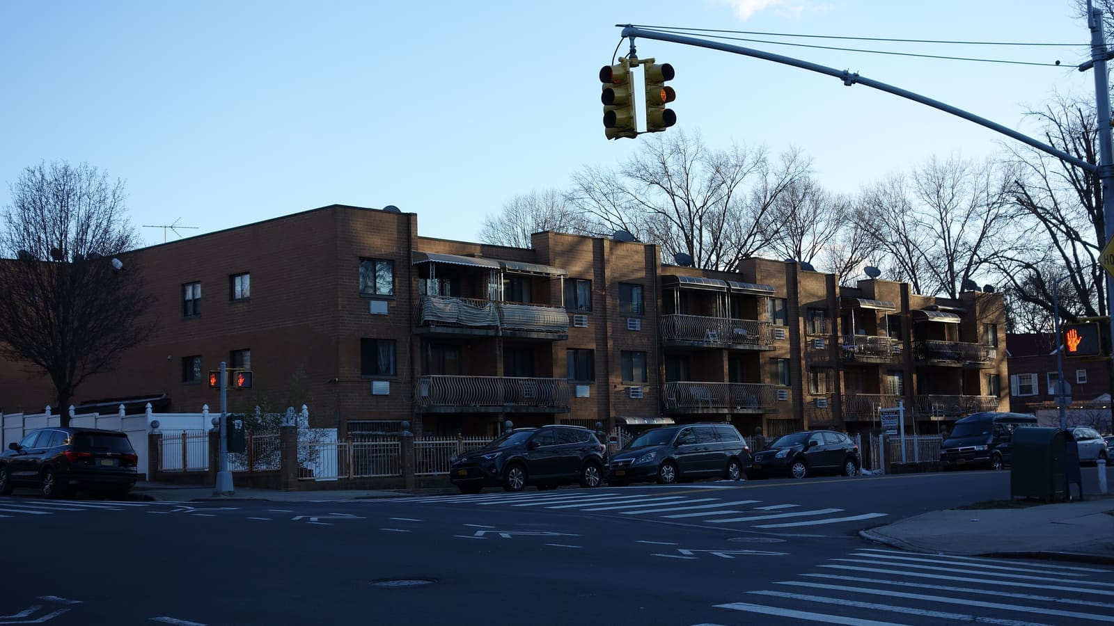 A three-story tan brick apartment building with curved balcony railings and awnings, seen across an intersection with cars parked in front.