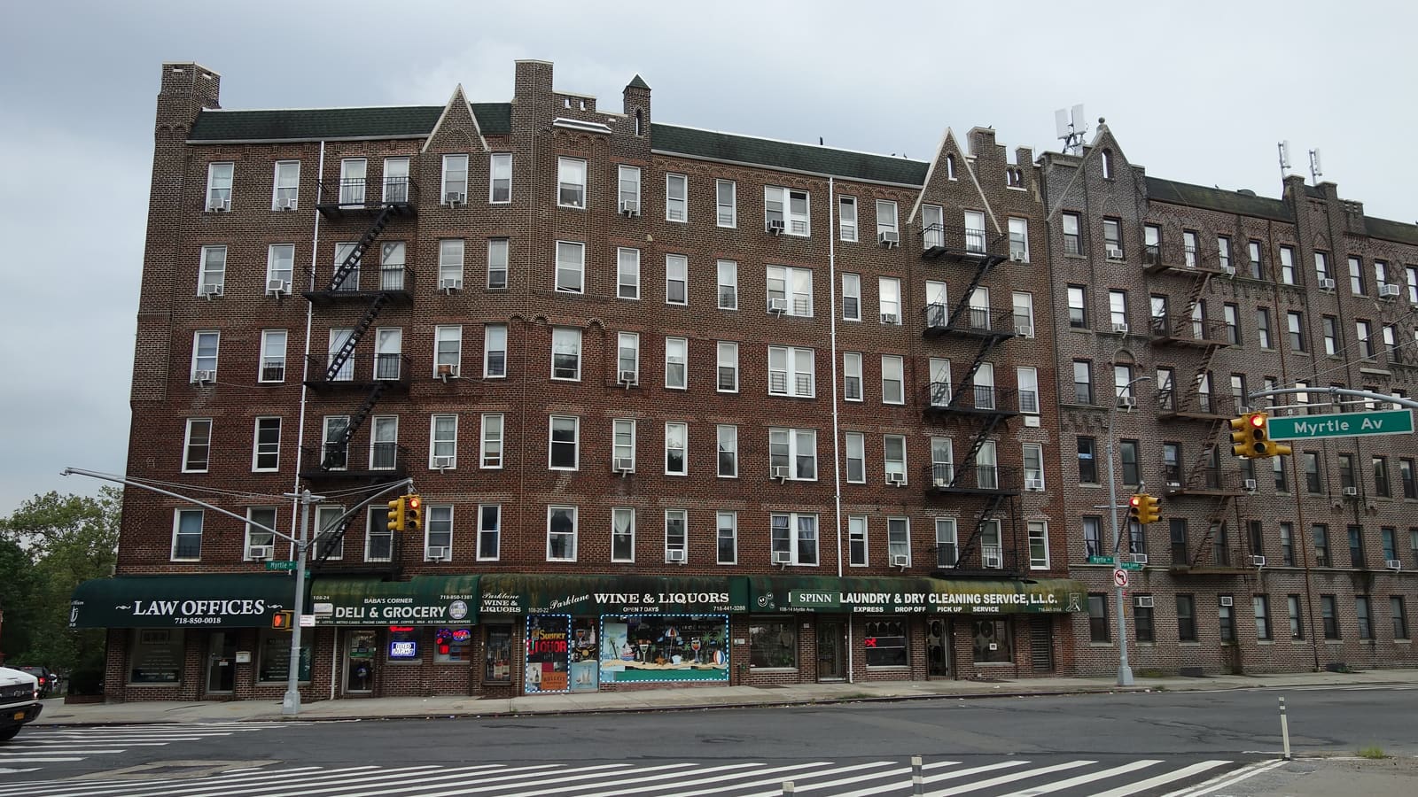 A row of six-story brick apartment buildings with fire escapes above ground-floor shops, with a Myrtle Av street sign at the right.