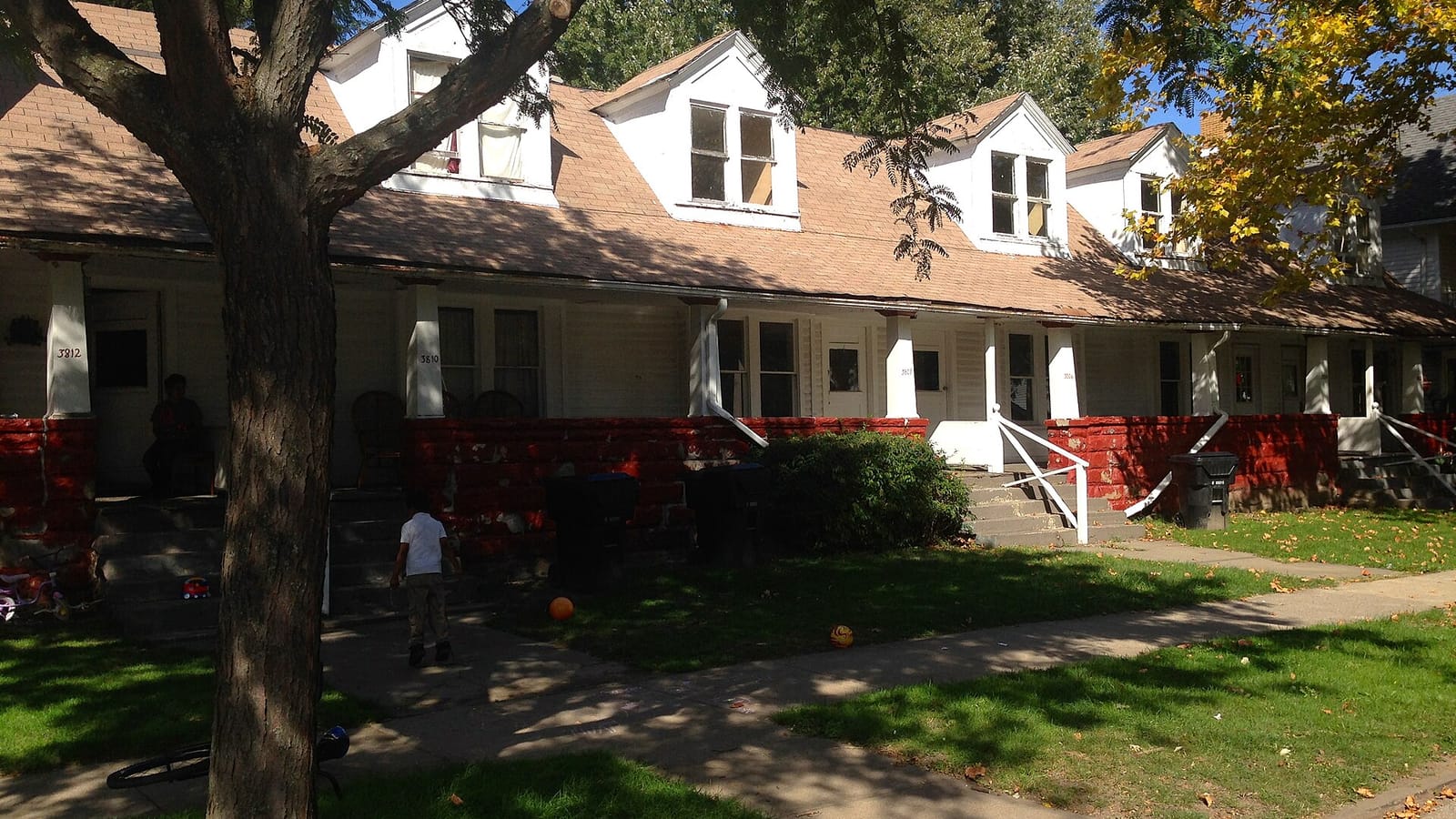 Low brick apartments with a long covered porch and front lawn beneath a tree