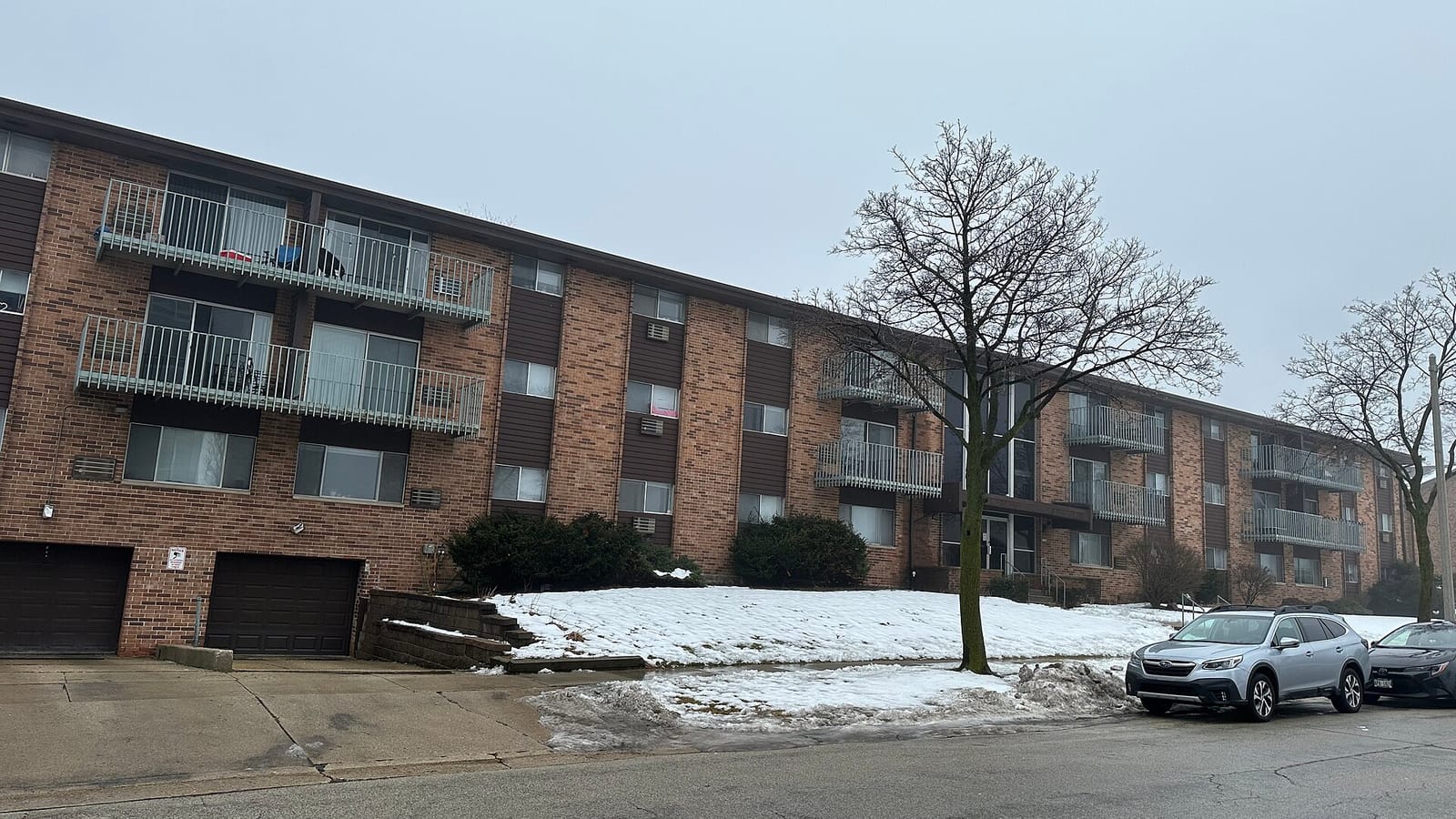 Low brick apartment block with balconies beside a sidewalk and patches of snow