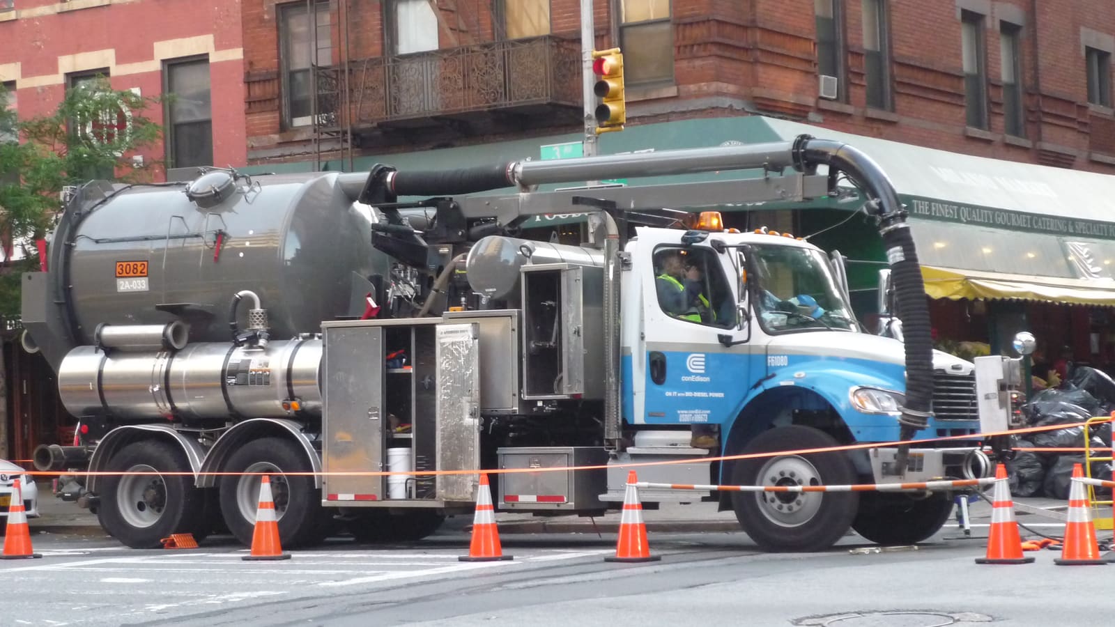 A blue and white Con Edison utility truck with a large vacuum tank parked behind orange traffic cones at a street corner, with brick apartment buildings behind it