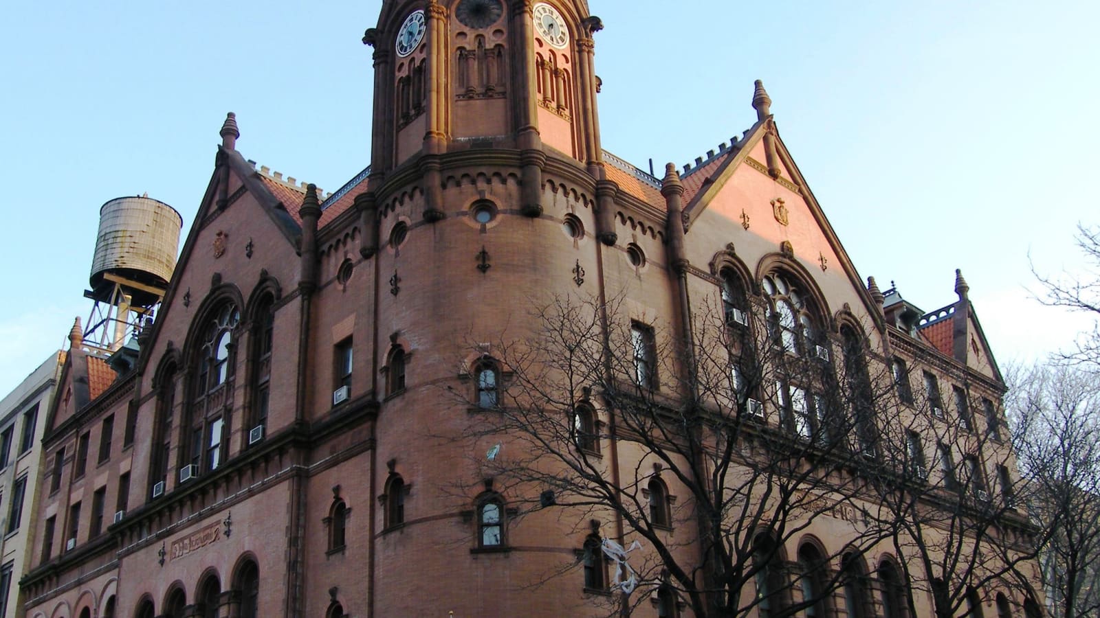A red brick and terracotta courthouse with a round corner tower, arched windows, gabled roofs and a clock tower, seen from the street through bare tree branches