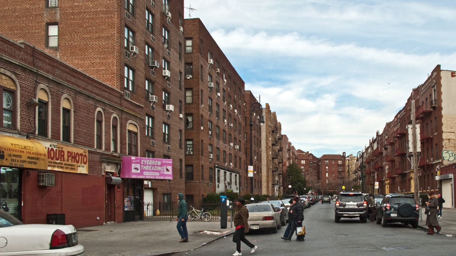 A street lined with six-story brick apartment buildings with fire escapes and window air conditioners, parked cars along both curbs and a few people crossing at the corner