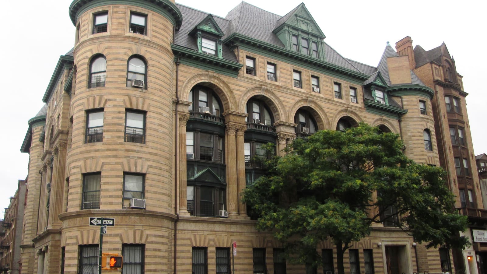 A tan brick and stone apartment building with round corner towers, tall arched windows, green copper trim and window air conditioners, behind a street tree.