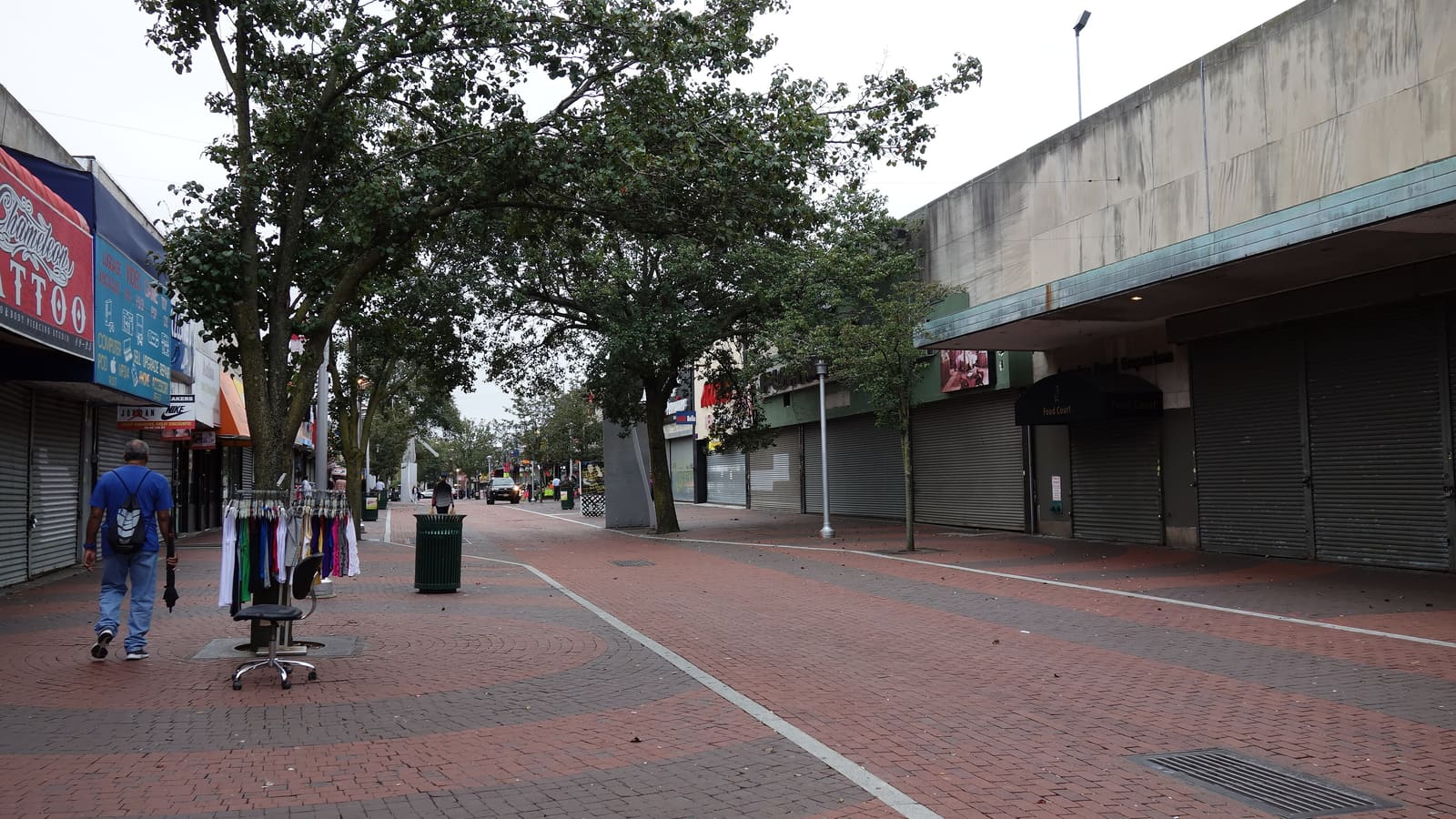 A brick-paved pedestrian street lined with trees and shops with their shutters down, a rack of clothes on the pavement and a man walking away from the camera.