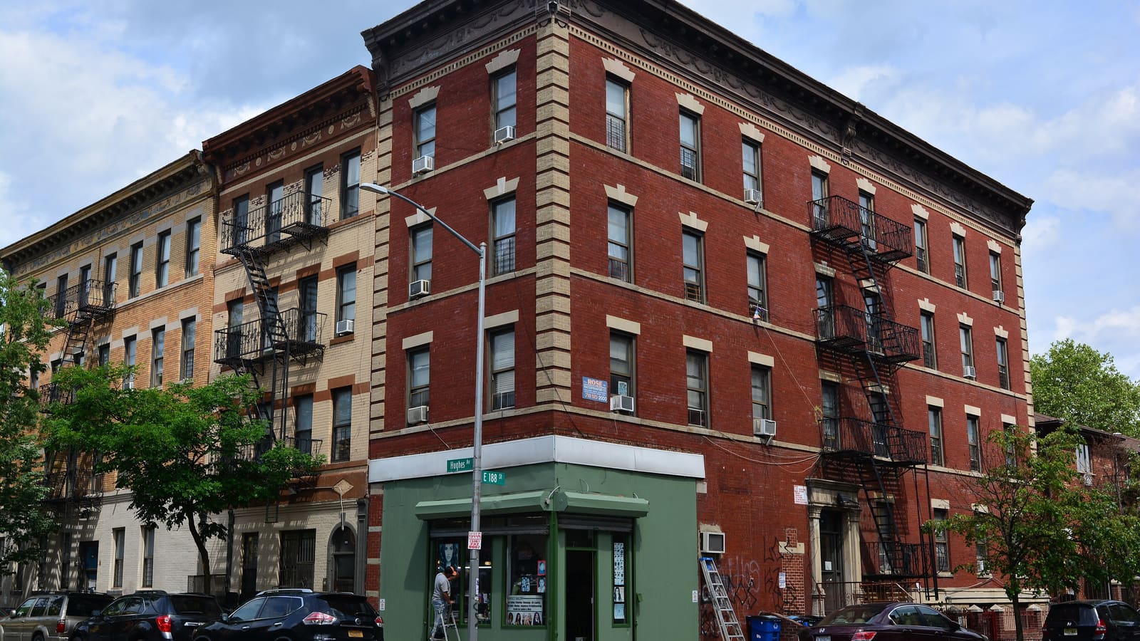 A four-story red brick corner apartment building with fire escapes and a small green storefront at street level, next to tan brick walk-up buildings.