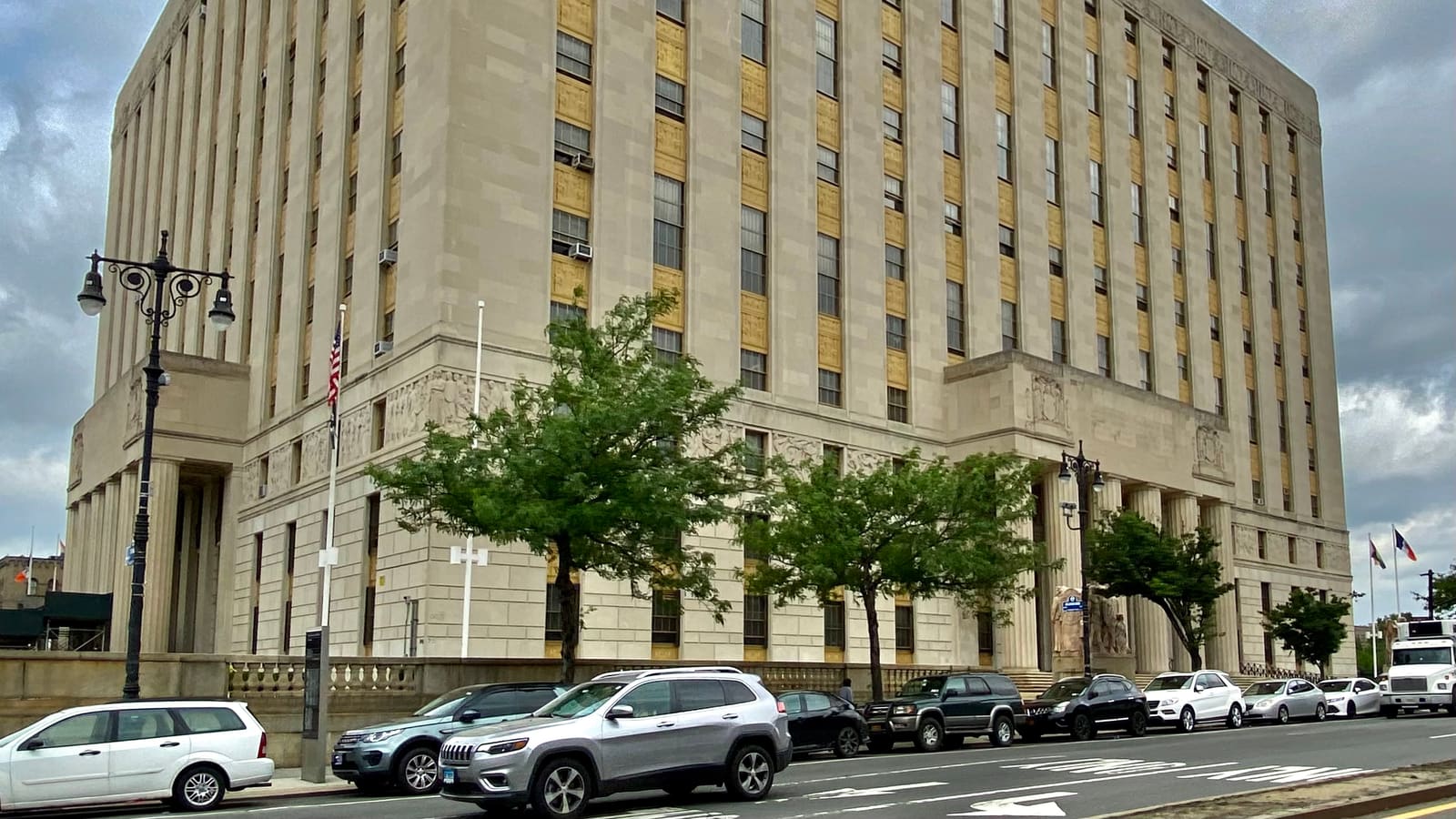 A large limestone courthouse with tall window bays and columned entrances, with street trees and parked cars along the road in front.