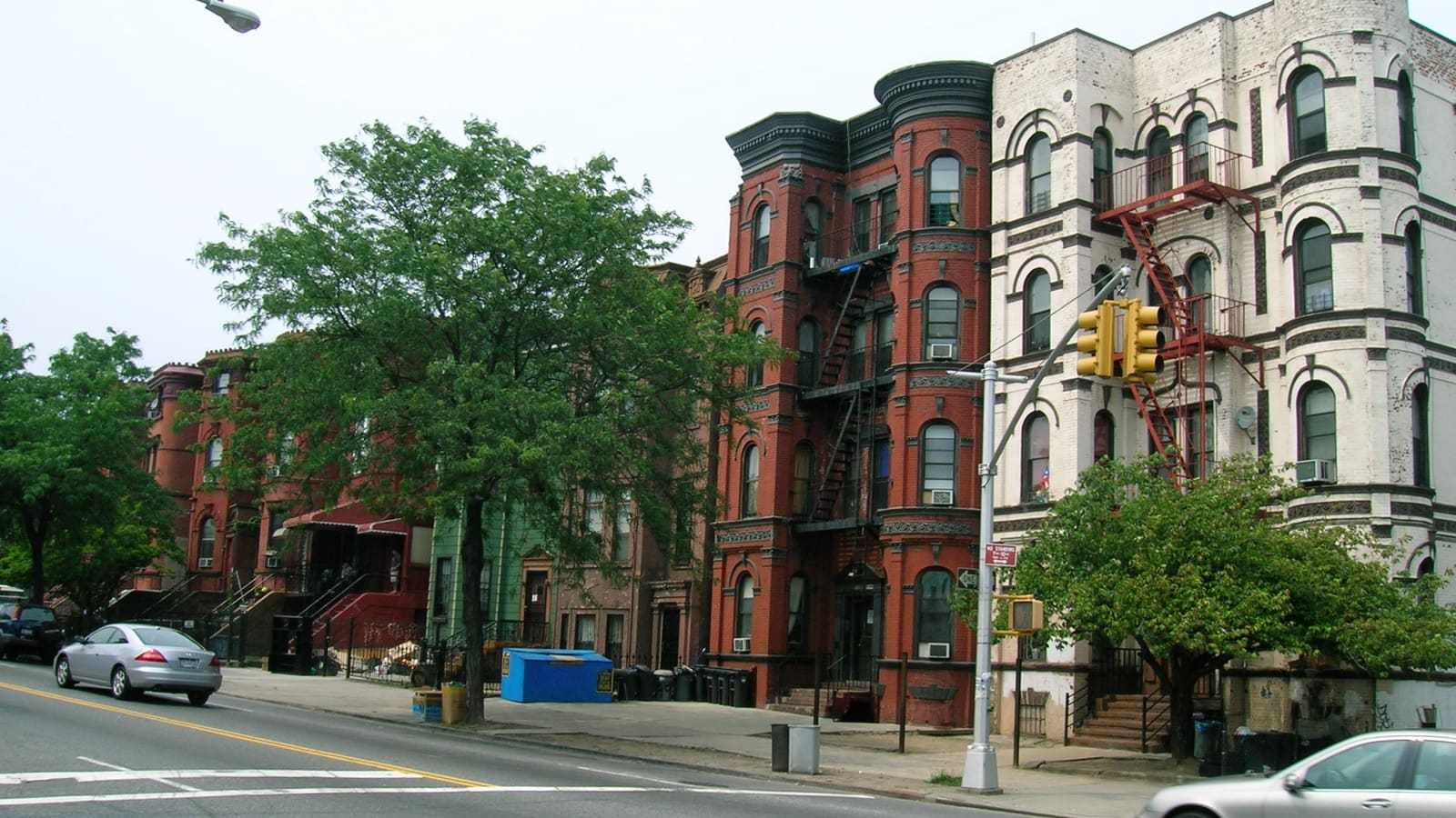 A street of four-story red and white brick apartment buildings with rounded bays and fire escapes, beside brownstones with stoops, street trees and a traffic light.