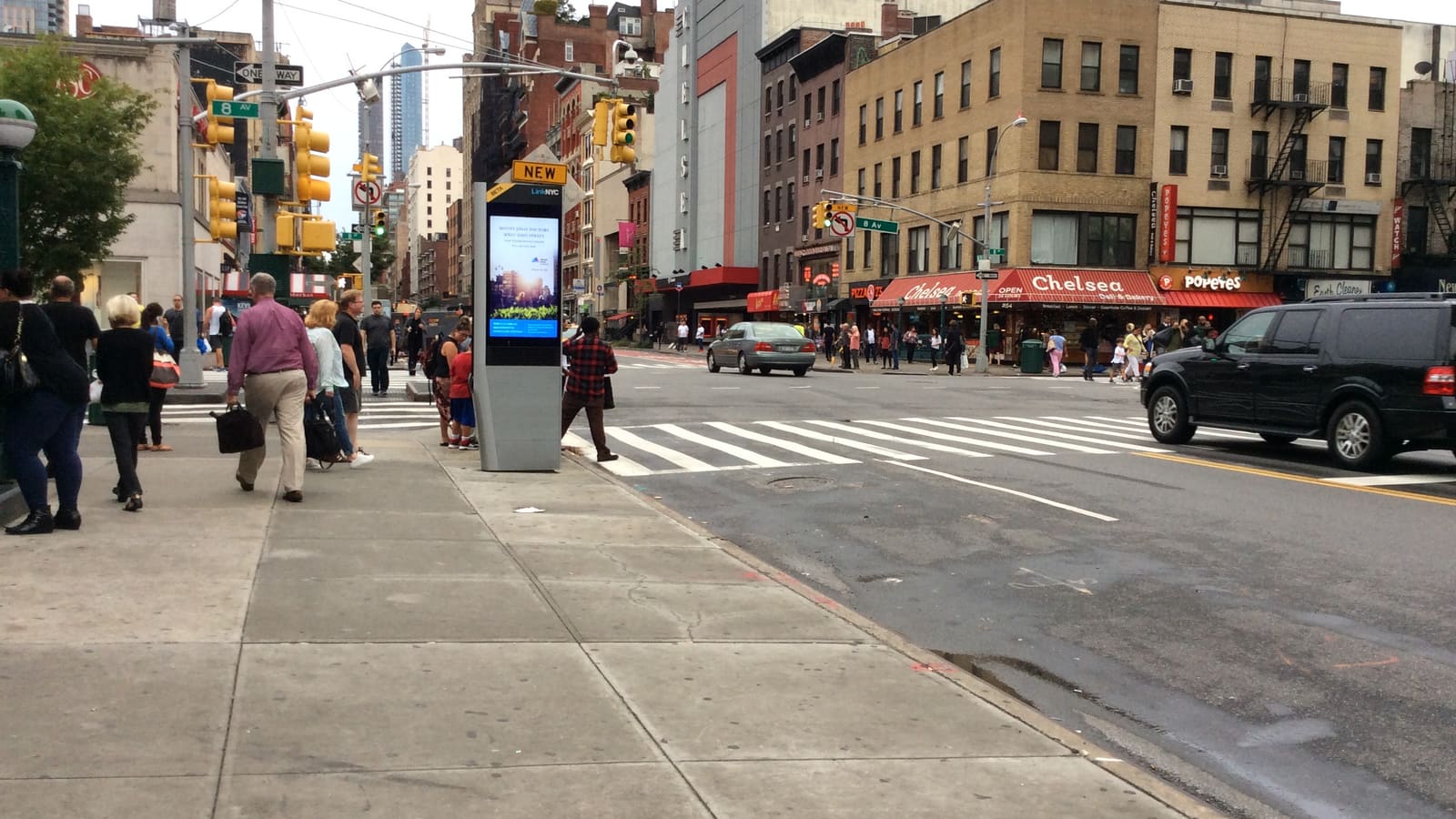 A tall grey LinkNYC kiosk with a lit screen on a wide sidewalk beside a crosswalk, with an 8 Av street sign, traffic lights, people walking away from the camera and storefronts across the avenue.