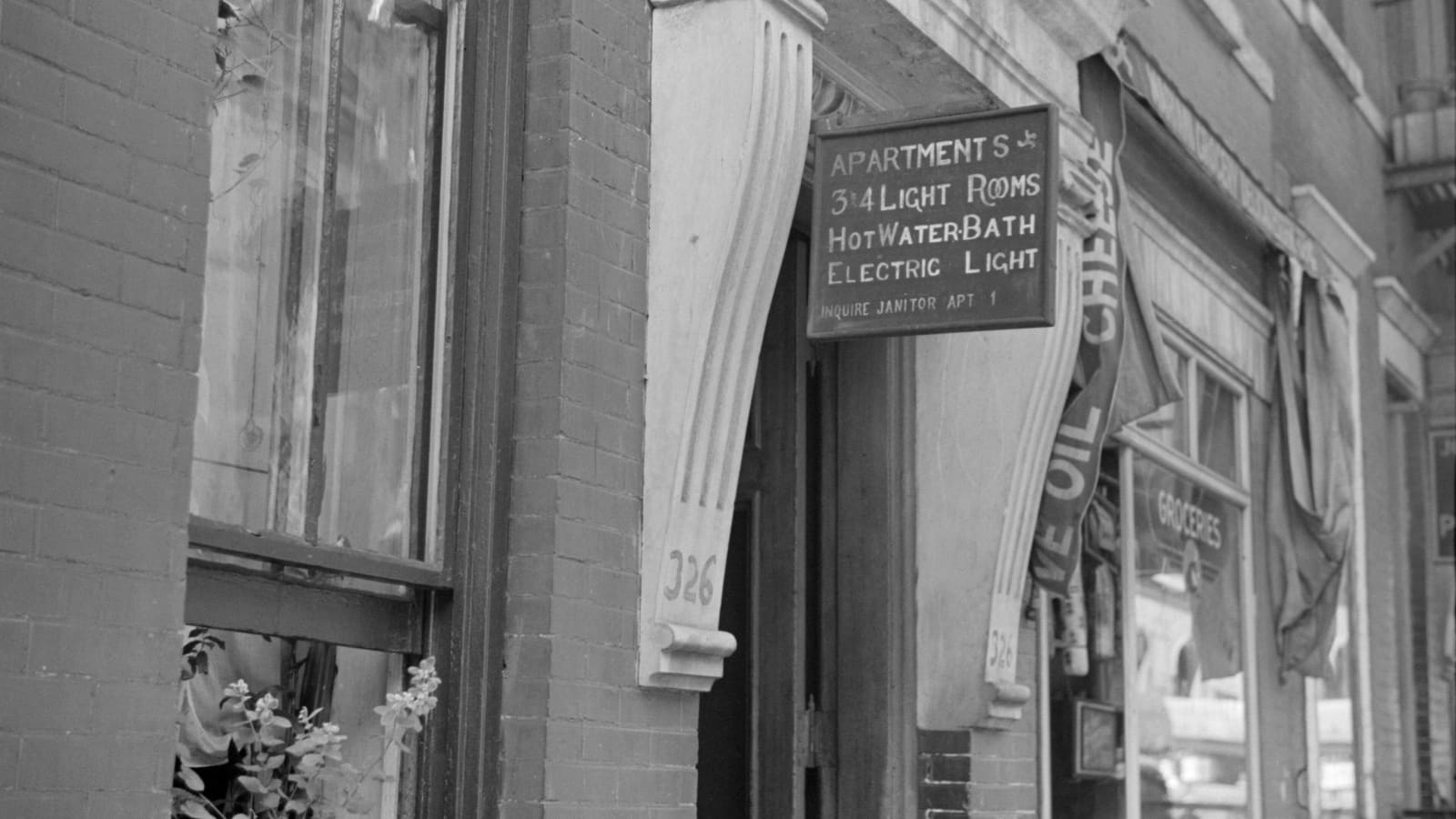Black-and-white photograph of a hand-lettered sign over a brick building's doorway reading Apartments, 3 and 4 light rooms, hot water, bath, electric light, inquire janitor apartment 1