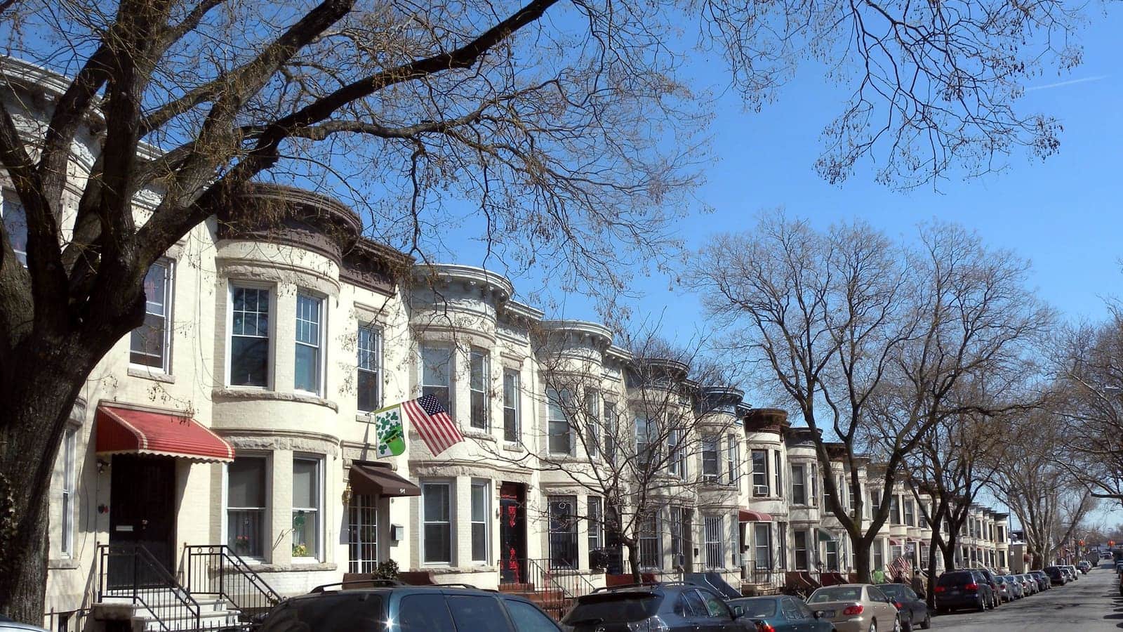 A long row of two-story cream-colored rowhouses with rounded bay fronts on a street lined with bare trees and parked cars, under a blue sky