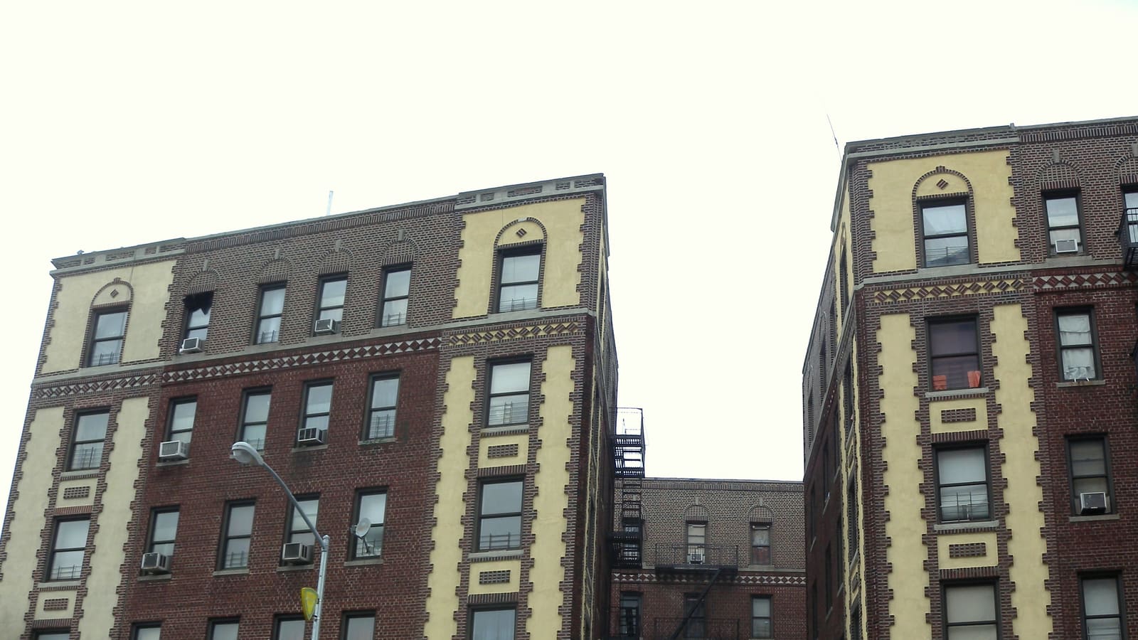 Upper floors of two six-story red-brick apartment buildings with cream panels, window air conditioners and a fire escape in the gap between them, under an overcast sky