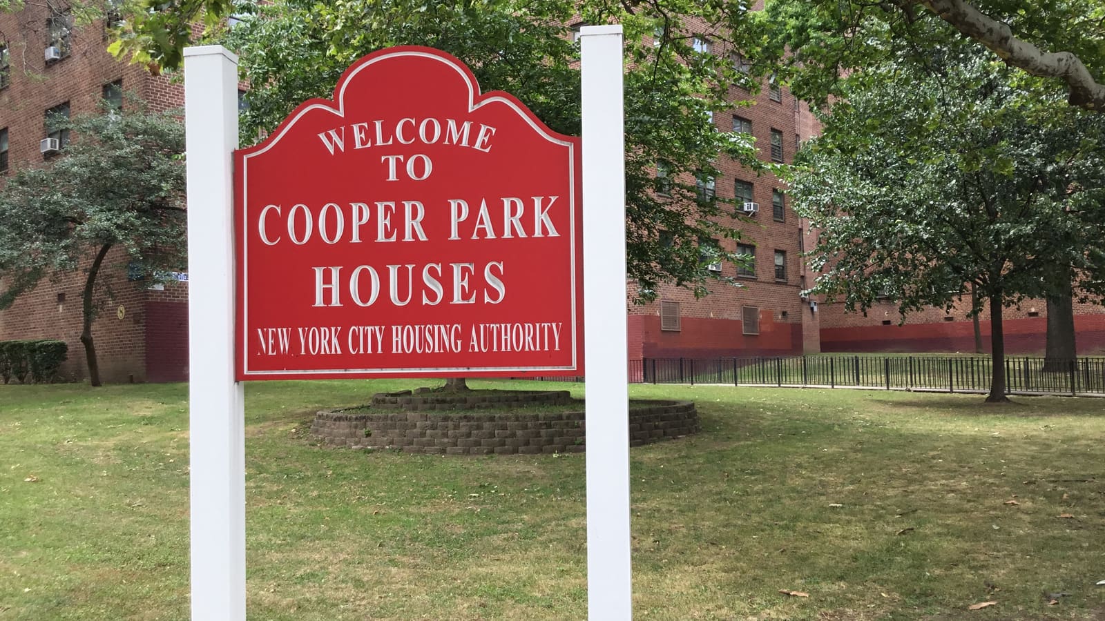A red sign reading Welcome to Cooper Park Houses, New York City Housing Authority, on a lawn in front of brick apartment buildings and trees