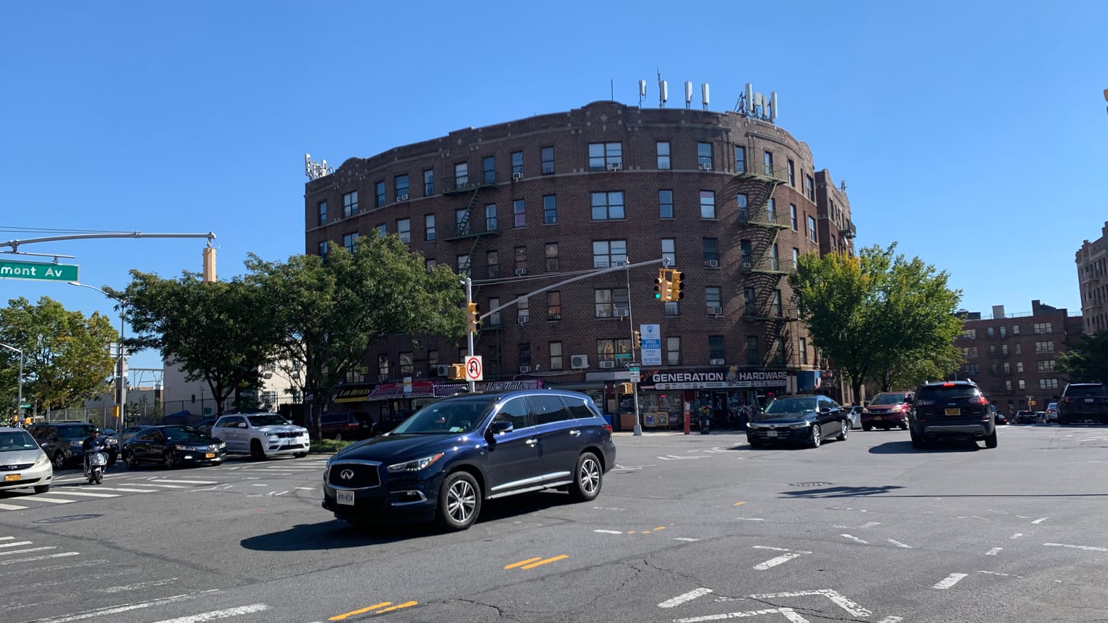 A six-story brick apartment building with fire escapes and ground-floor shops on a street corner, with cars crossing the intersection in front