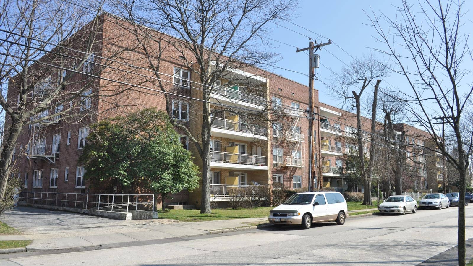 A four-story red brick apartment building with balconies on a tree-lined residential street, with cars parked at the curb