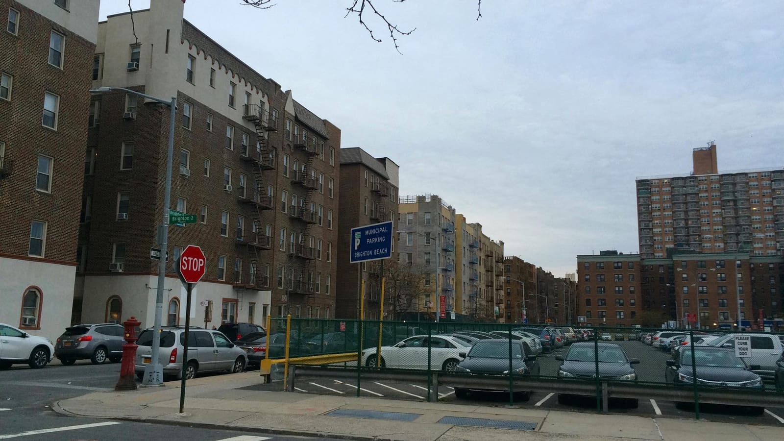 A row of six-story brick apartment buildings with fire escapes along a street, with a parking lot and a taller apartment tower to the right.