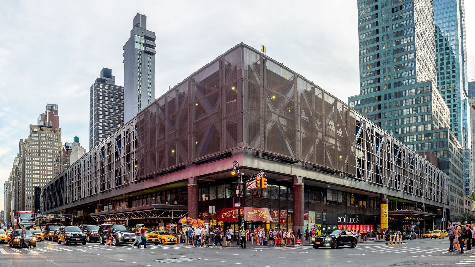 A large steel-framed bus terminal on a city street corner with taxis, cars and people crossing in front and towers behind it.