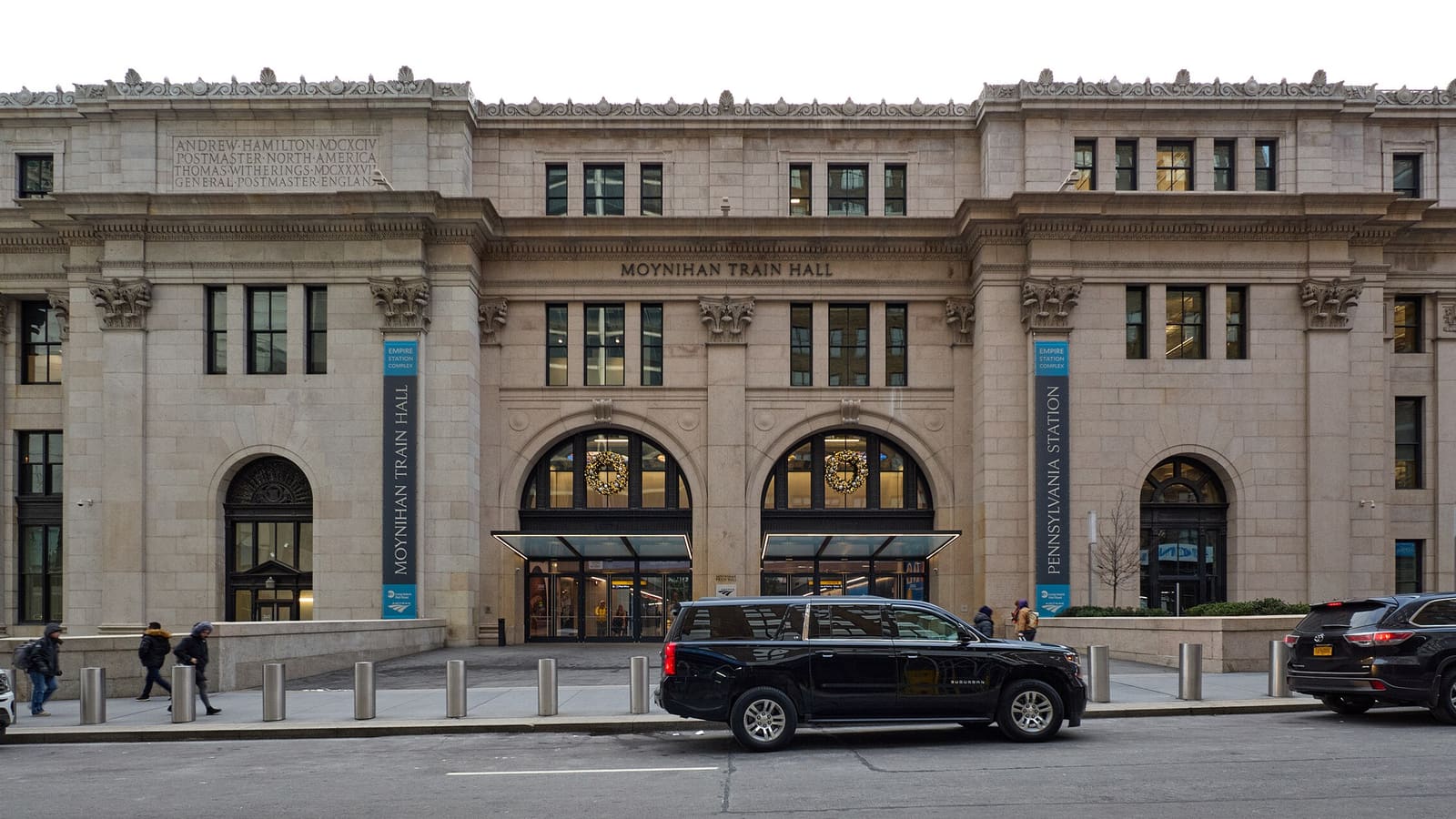 A wide stone building with two arched glass entrances, the words Moynihan Train Hall carved above them, and banners reading Moynihan Train Hall and Pennsylvania Station on either side.