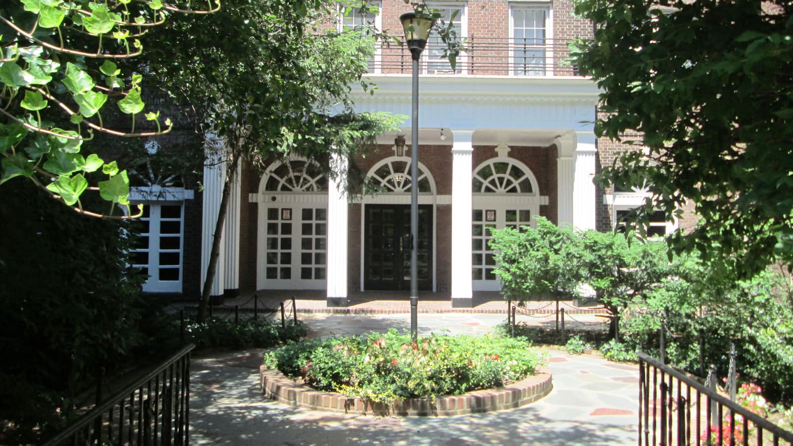 A brick apartment building entrance with white columns and arched fanlight windows, seen across a paved garden court with a lamp post and a round planted bed.