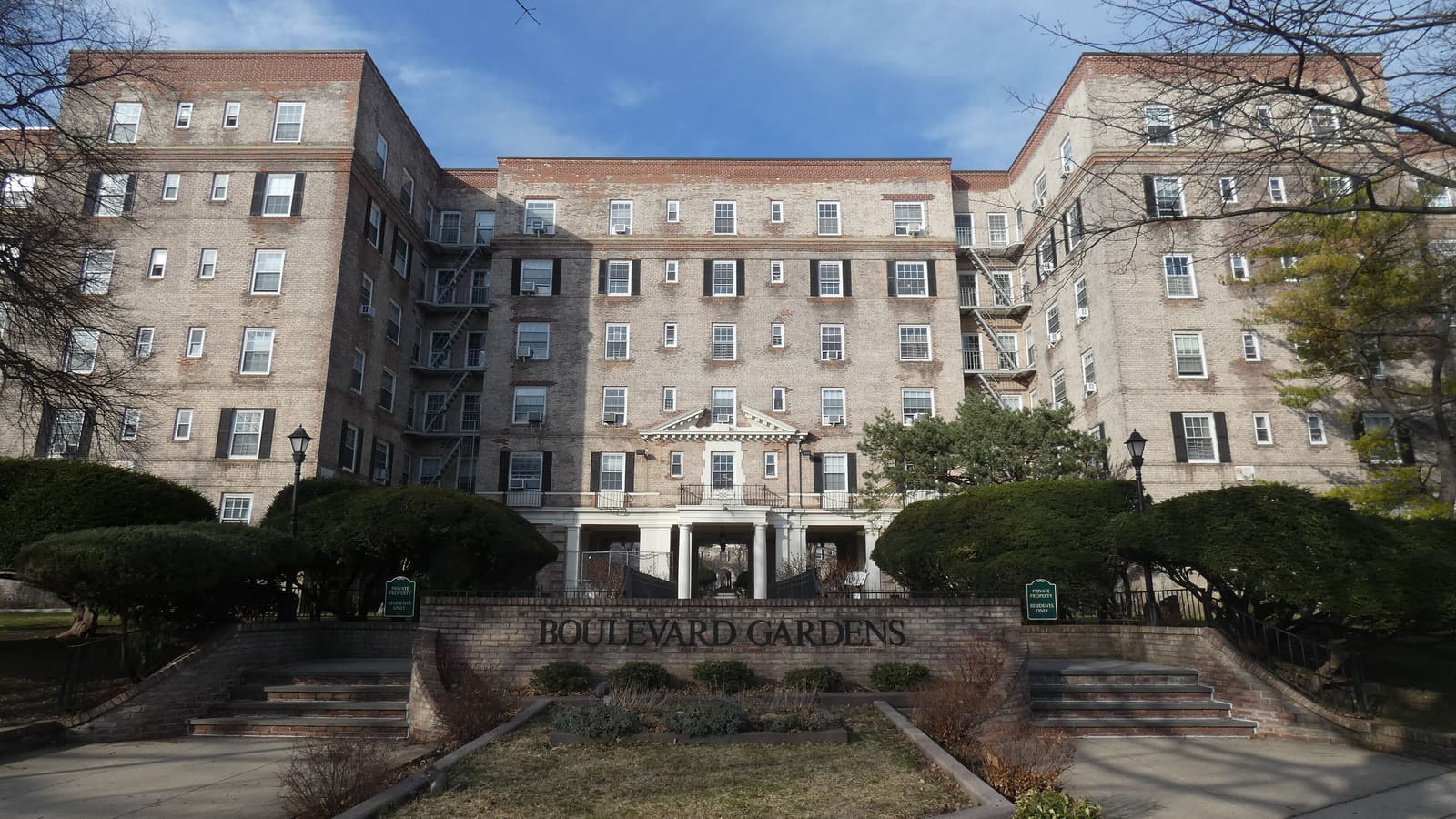 A six-story brick apartment building with fire escapes and white-columned entrance, behind clipped evergreens and a low brick wall lettered Boulevard Gardens.