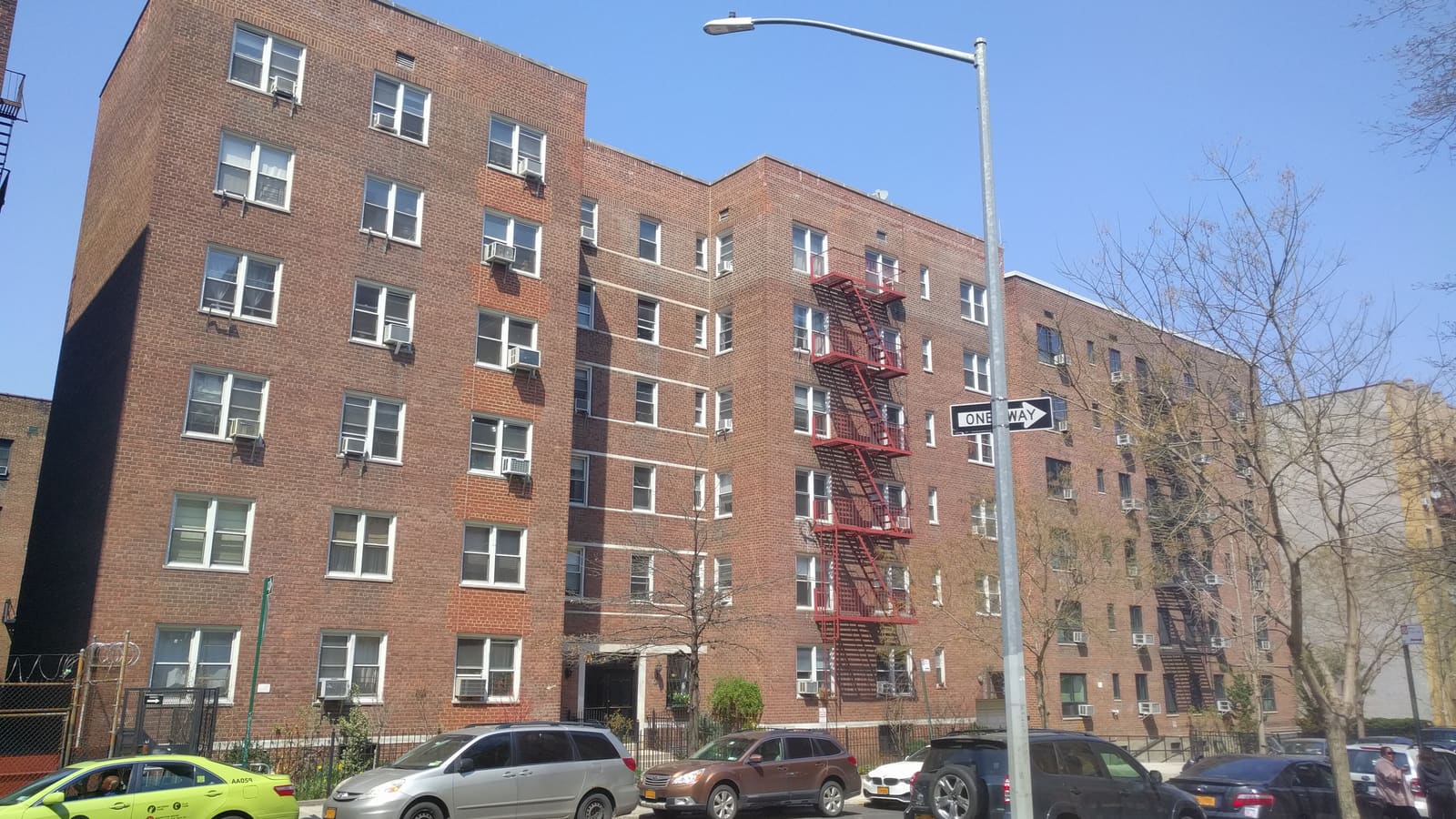 A six-story red brick apartment building with window air conditioners and a red fire escape, cars parked along the curb in front