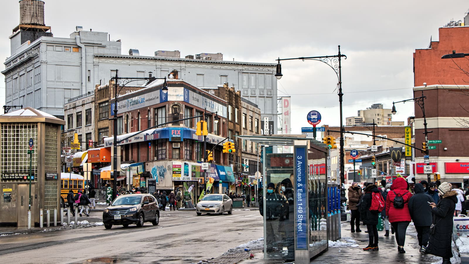 A busy intersection lined with shops and offices, with a bus shelter marked 3 Avenue and East 149 Street, a subway entrance, cars and people in winter coats waiting on a wet sidewalk
