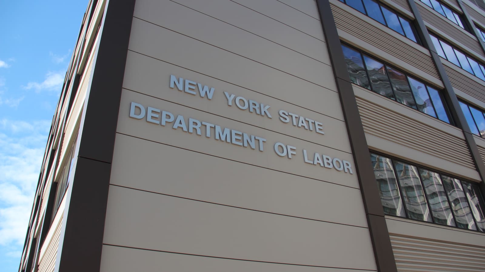 Metal letters reading New York State Department of Labor on the tan wall of an office building, seen from the street below against a blue sky
