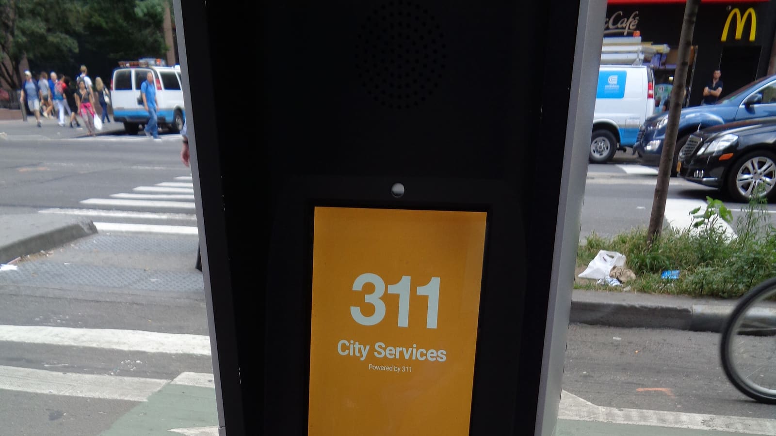 Close view of a street kiosk whose orange screen reads 311 City Services, with a crosswalk, parked vans and distant pedestrians behind it