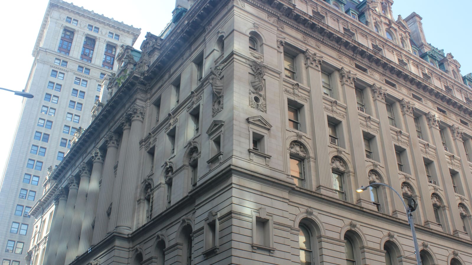 Looking up at the carved stone corner of a seven-story Beaux-Arts courthouse with columns and arched windows, a taller municipal tower behind it