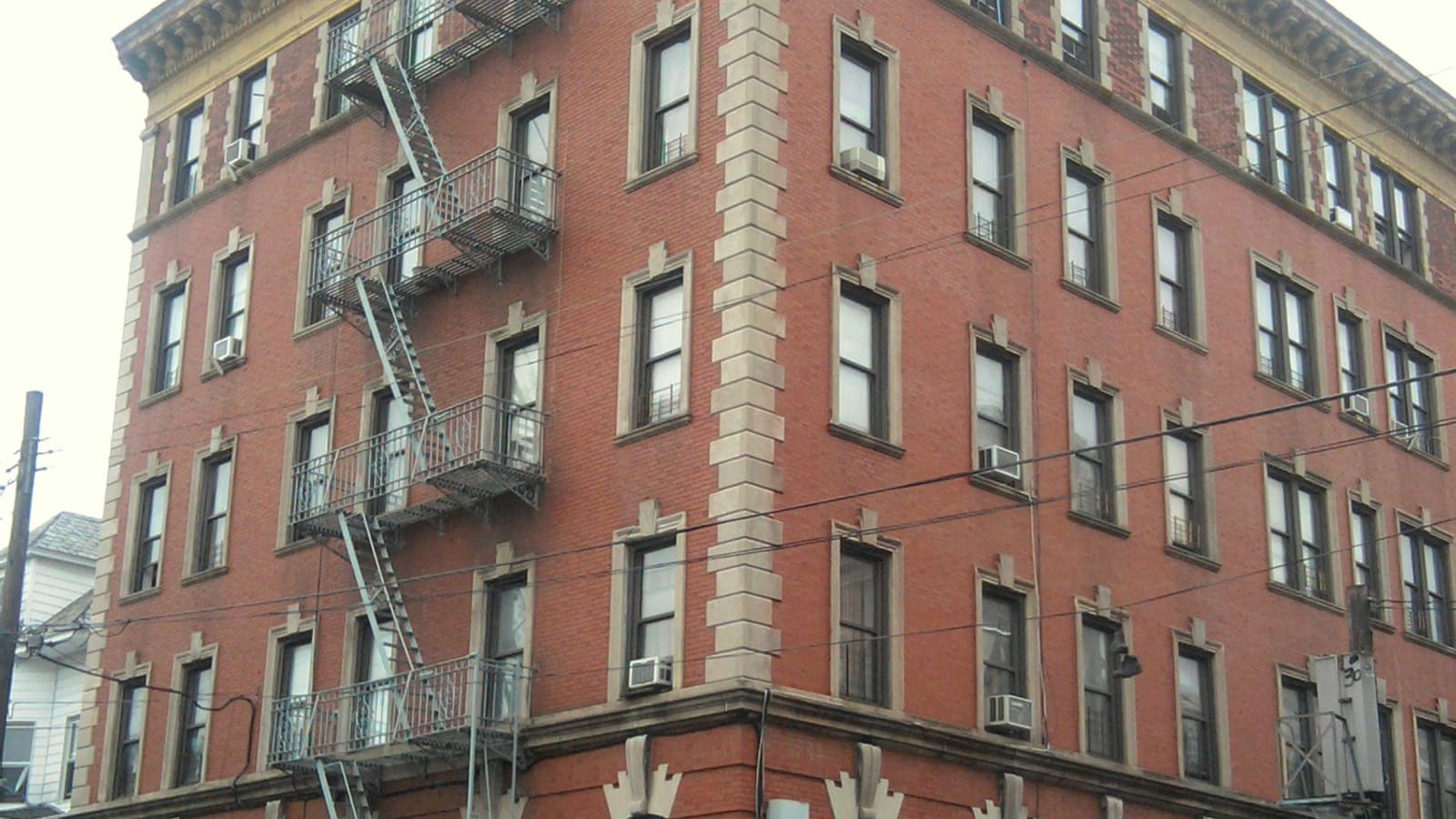 Corner of a five-story red-brick apartment house with stone quoins, a metal fire escape and window air conditioners, under an overcast sky