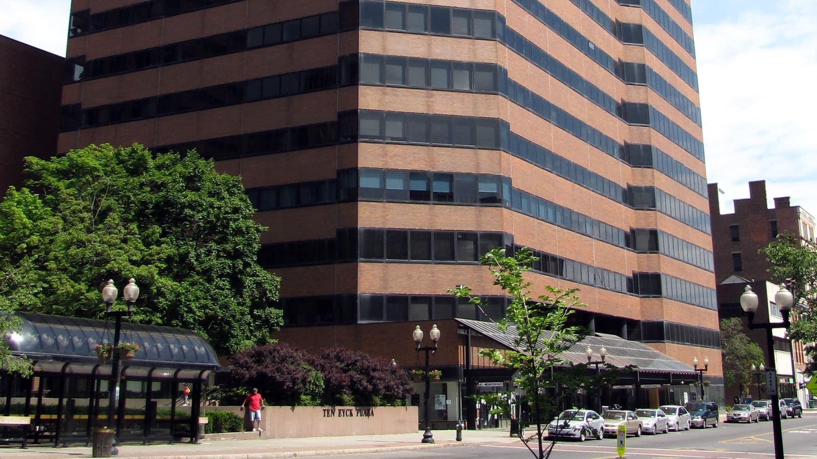 A brown brick office tower with bands of dark windows above a plaza with street lamps and parked cars.
