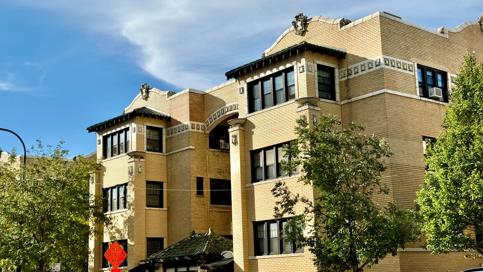 A three-storey buff-brick courtyard apartment building with green tile roofs over its bay windows, behind street trees under a blue sky