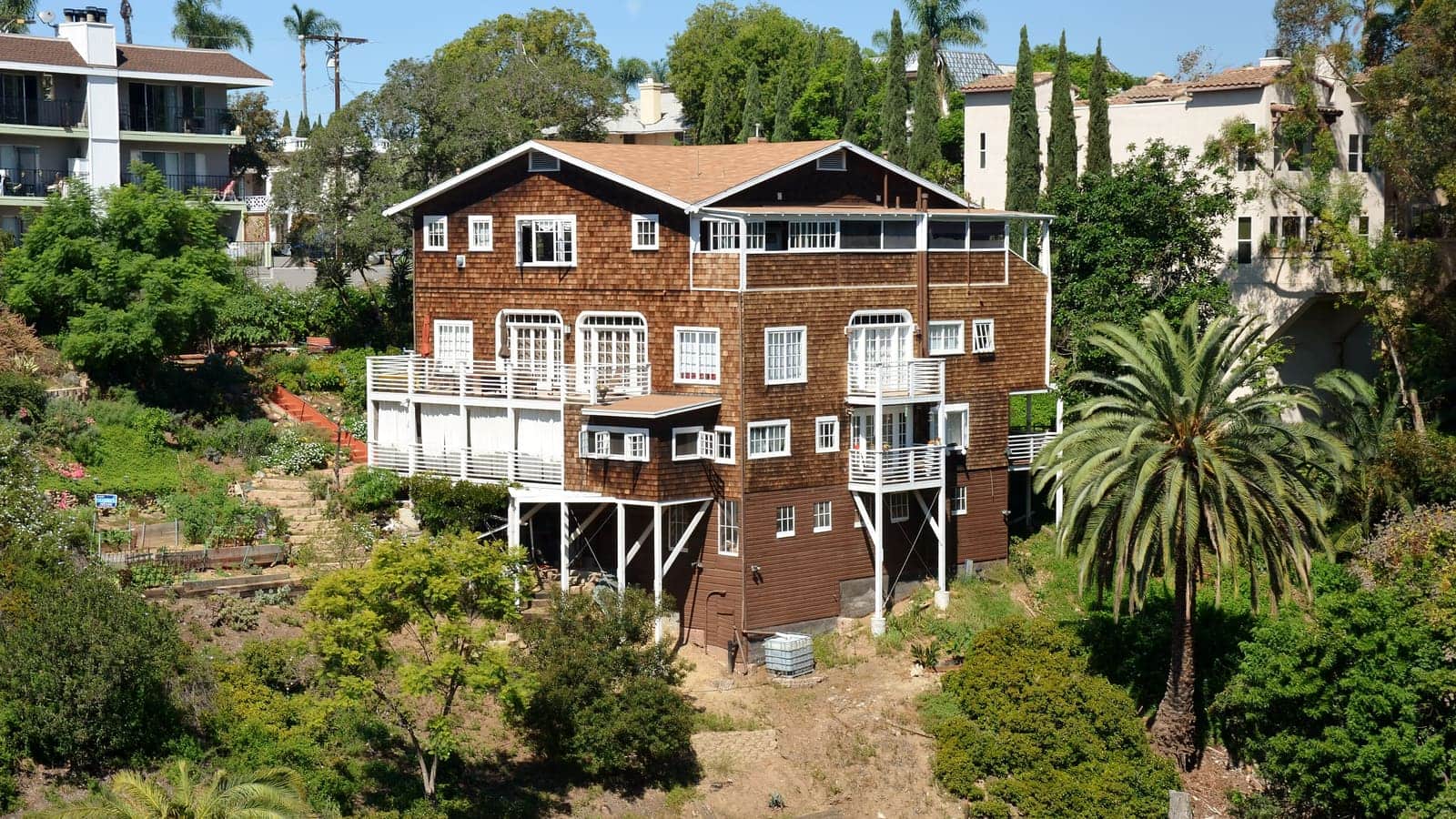 A three-storey brown-shingled residential building with white balconies on a planted canyon slope, with an apartment block and palms beside it