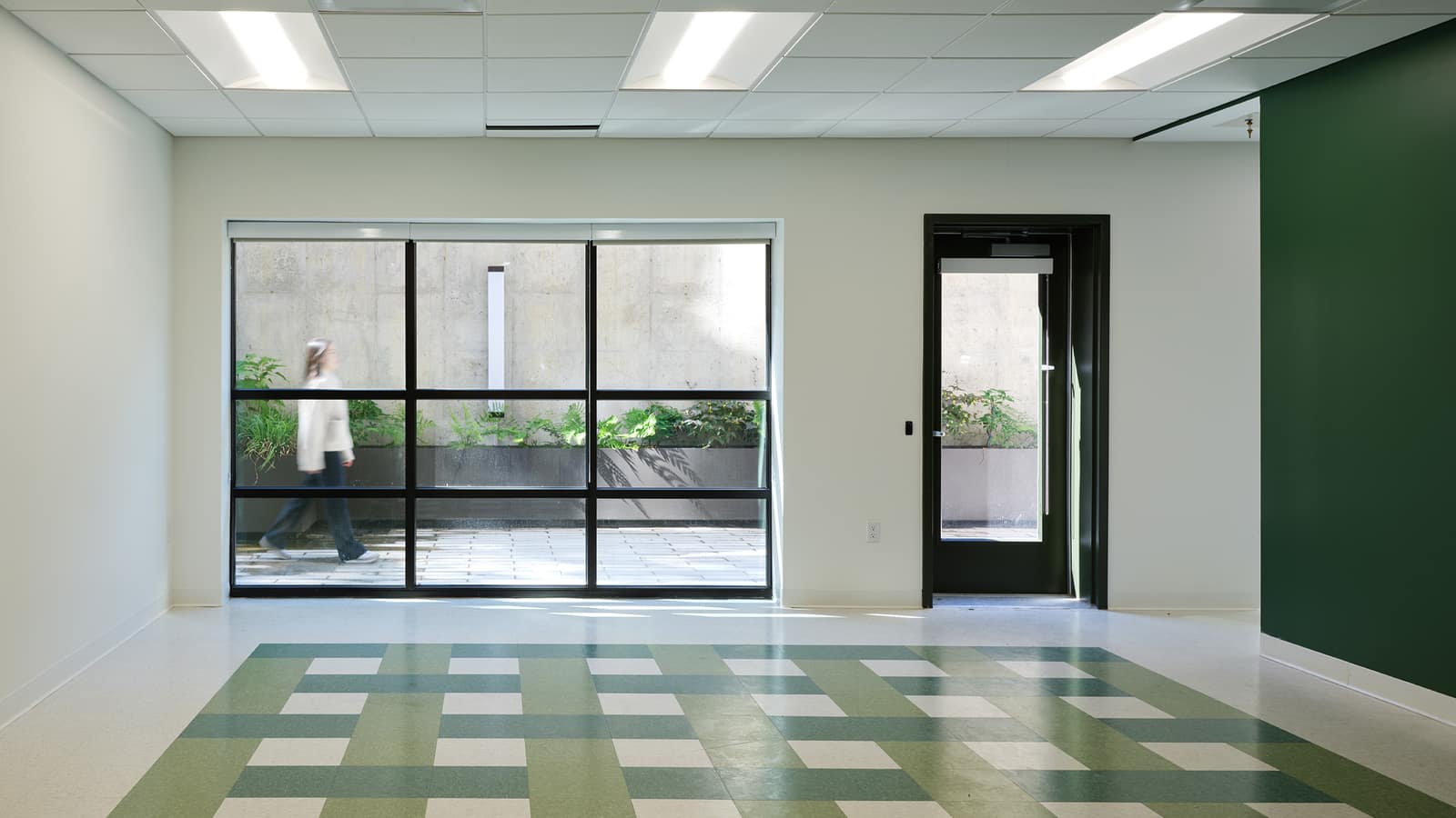 An empty room with a white tile ceiling and long lights, a green and white woven-pattern floor, a dark green accent wall, and a large window and glass door opening to a planted sunken courtyard where a blurred person walks past