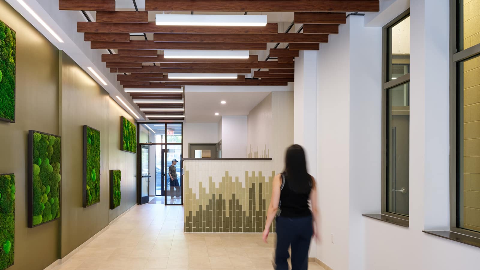 A lobby with wood slats hung across the ceiling, framed green moss panels on an olive wall, a front desk faced in green and cream tiles, a glass entry door at the far end and a person walking away from the camera