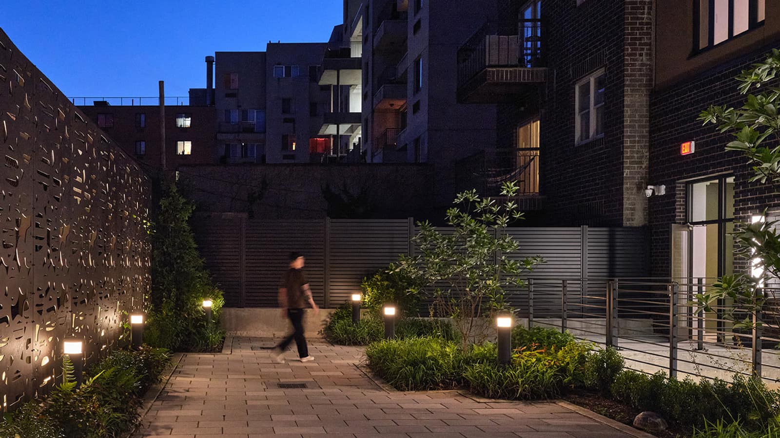 A rear yard at night with a paved path, low lit bollards, planted beds, a patterned metal screen wall on the left, a slatted fence at the back and a blurred person walking across