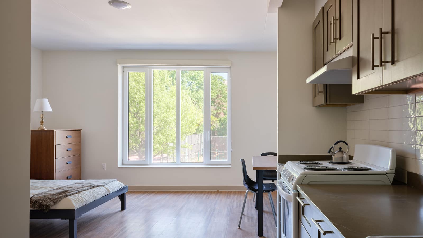 A studio apartment with a wood-look floor, a bed and a wooden dresser by a wide window looking onto trees, a small table with two chairs, and a kitchen wall with brown cabinets, a white tile backsplash and a white electric coil stove with a kettle
