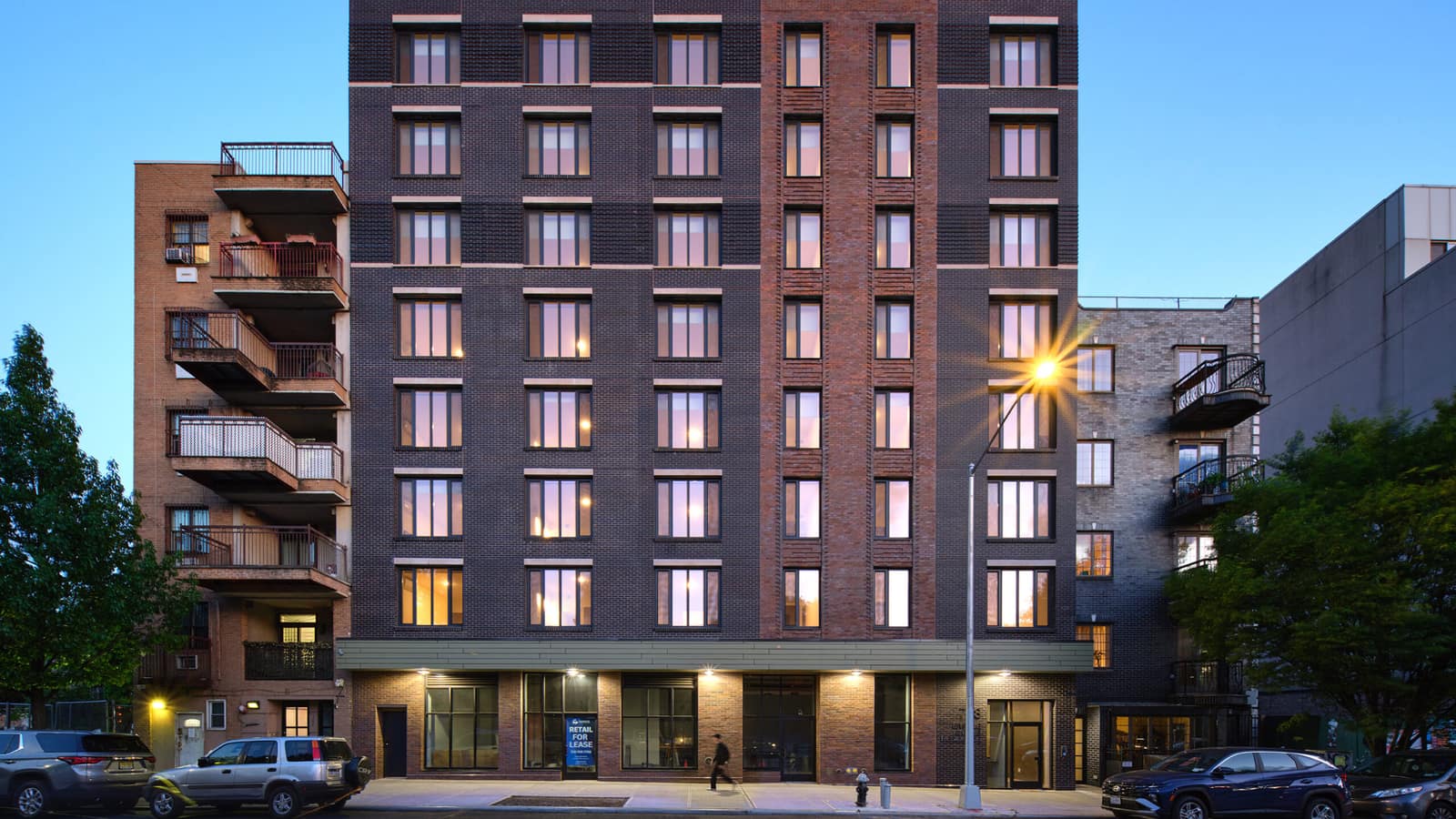 A nine-story apartment building of dark brown brick with a lighter red brick band running up the middle, rows of lit windows at dusk, a green metal band over glass storefronts, a Retail For Lease sign in one window, a lit street lamp, parked cars and a single walker on the sidewalk