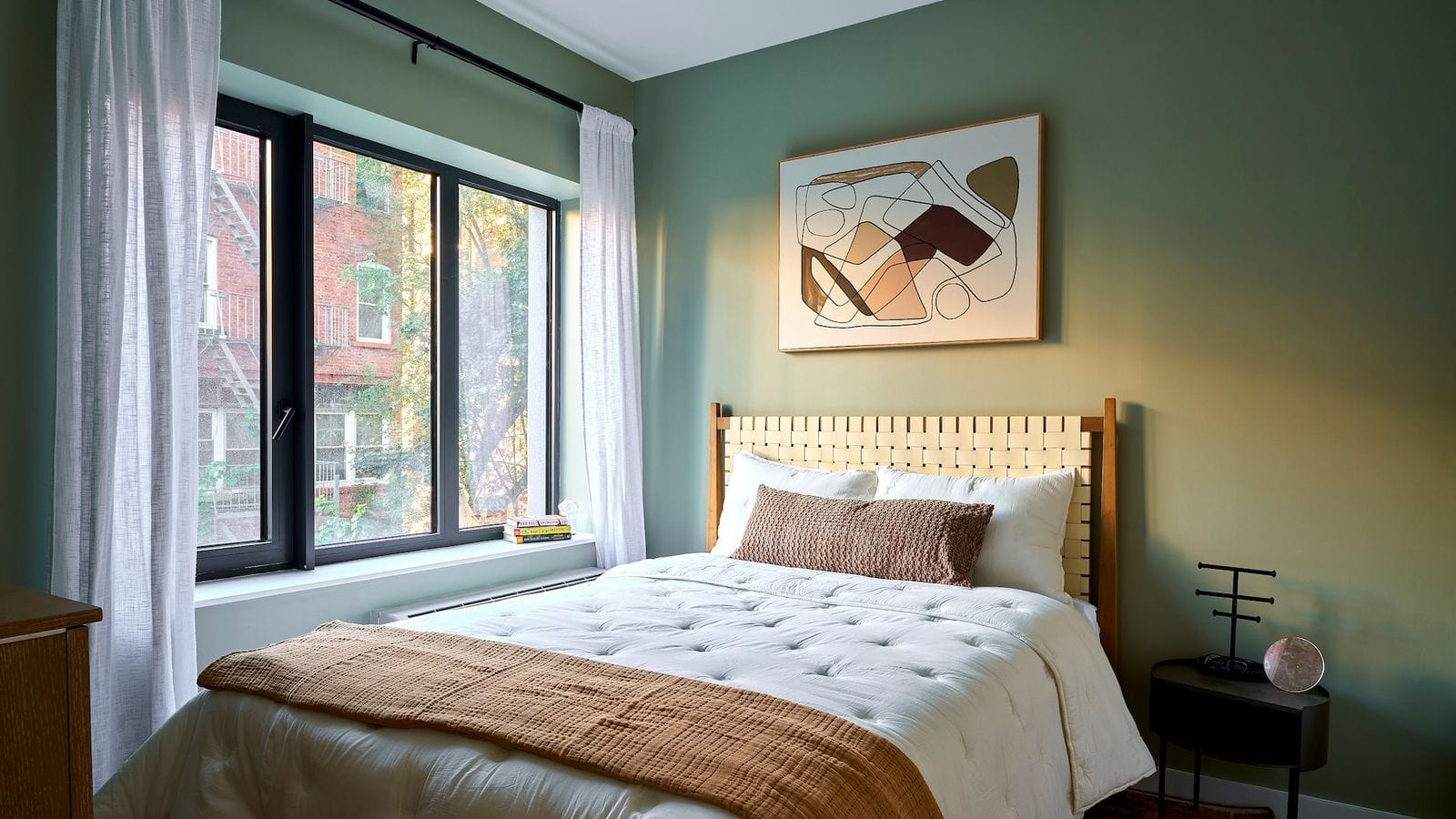Bedroom with a green wall, a large black-framed window with sheer white curtains looking onto a brick building with fire escapes and trees, a bed with a woven headboard and white and tan bedding, an abstract framed print and a small black side table