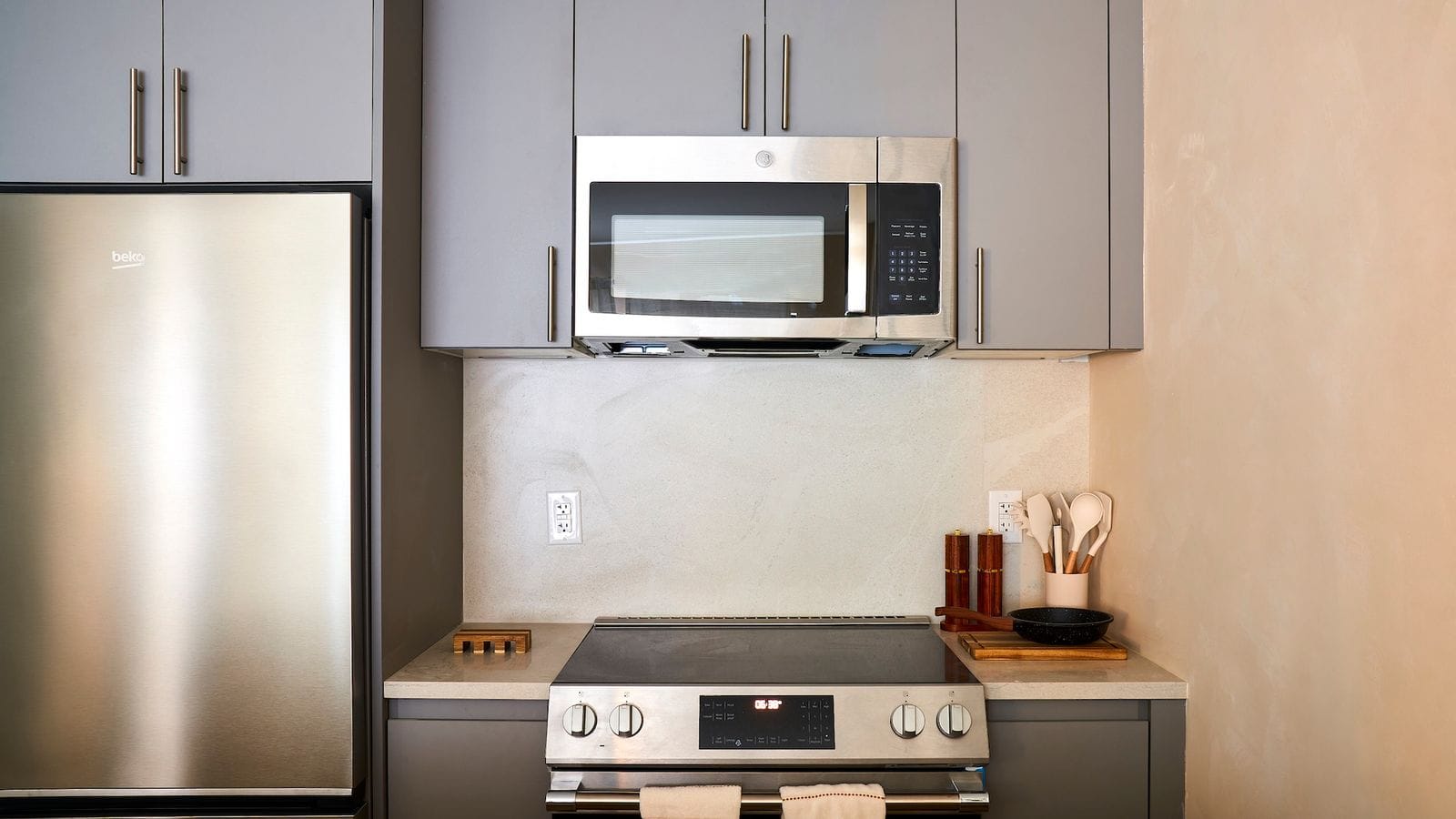 Kitchen wall with gray cabinets, a stainless-steel Beko refrigerator at left, a GE microwave above a smooth-top electric range with front knobs, a light stone backsplash and counter, and a small pan and utensils beside the range