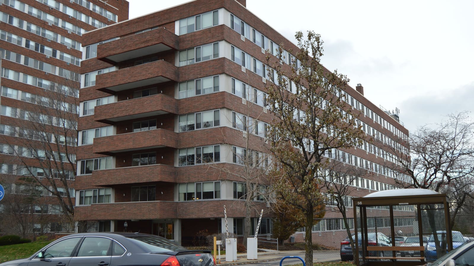 A six-story red brick apartment building with stacked balconies, a taller block behind it, and a bus shelter and parked cars in front