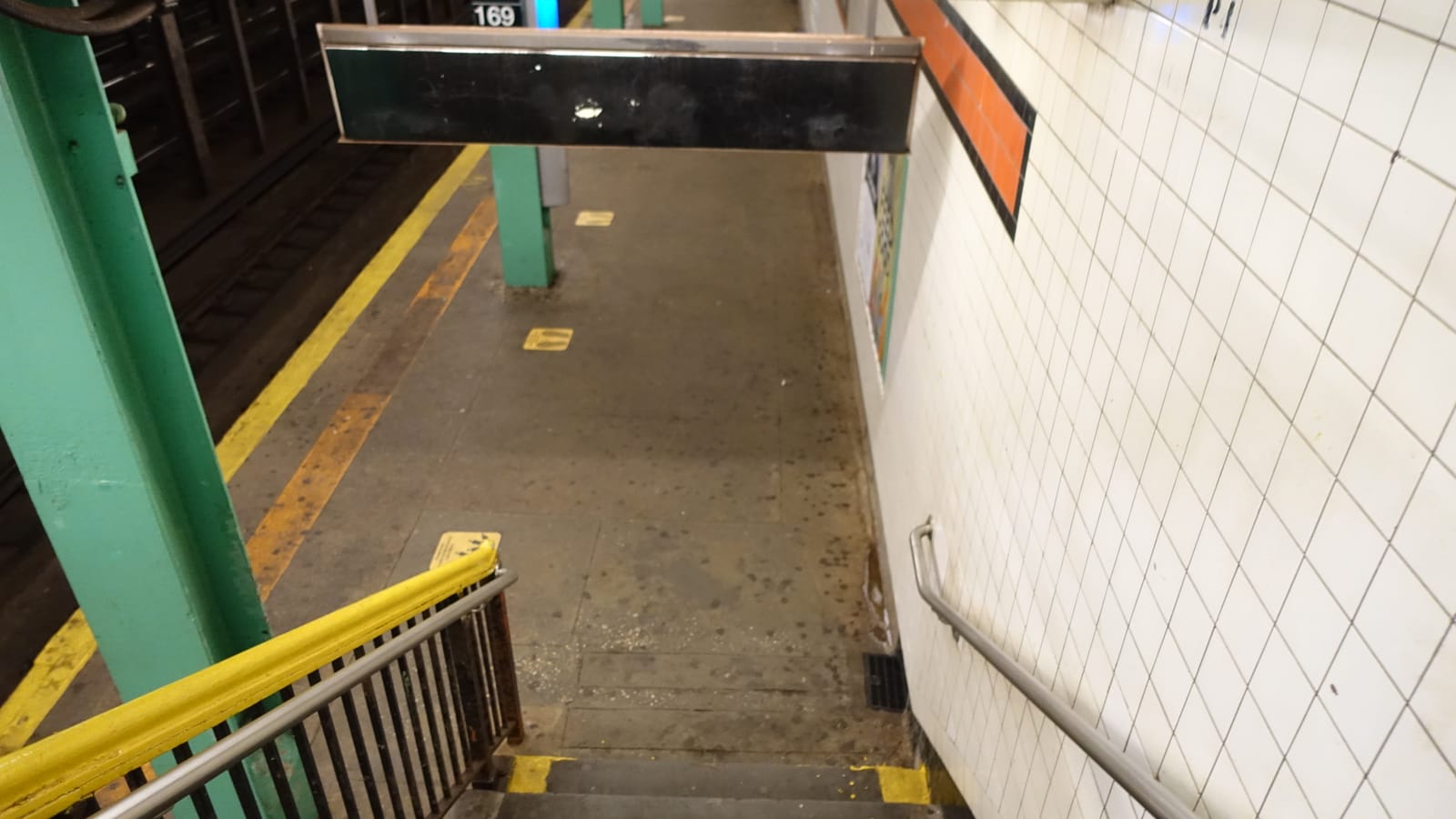 Looking down a stair with a yellow handrail onto an empty subway platform with green columns, a yellow edge strip, white tile walls with an orange band and a sign reading 169