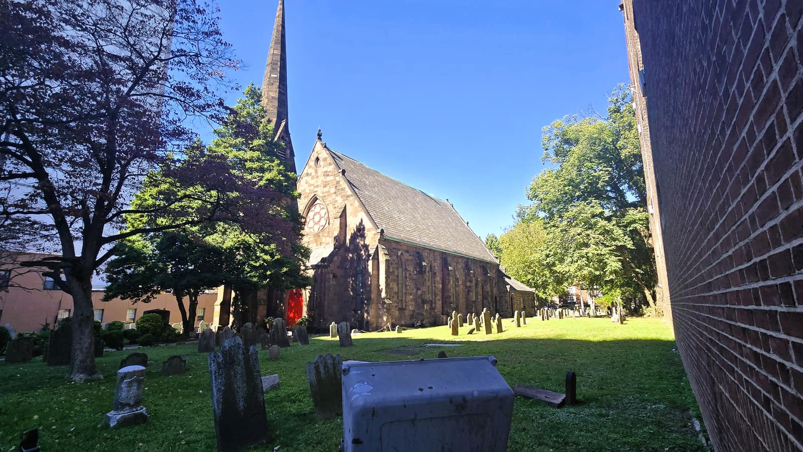 A brownstone Gothic church with a tall spire and a red door, standing in a grassy churchyard with old gravestones and trees under a blue sky, a brick wall at right