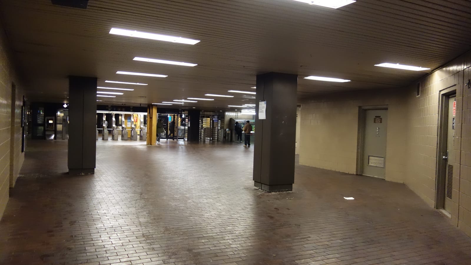 A wide underground subway mezzanine with a brown brick floor, dark square columns and tan tile walls, with turnstiles and a few distant figures at the far end