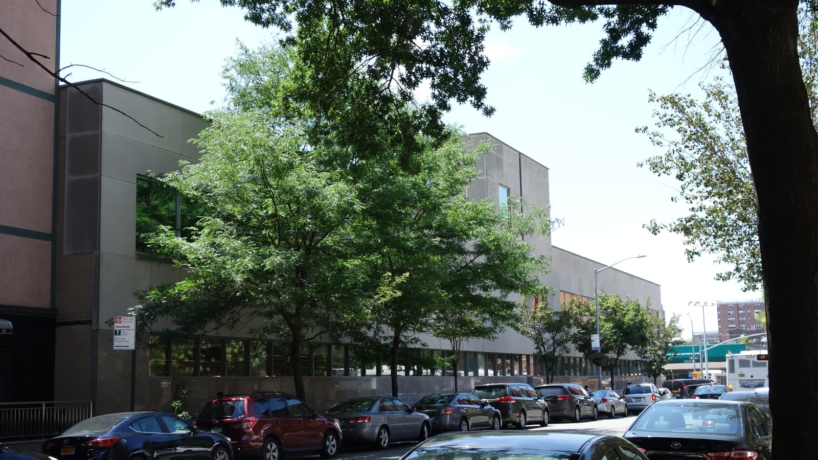 A long two-story beige library building with a tall window band behind a row of street trees, cars parked along the curb, a street sign at left and a city bus in the distance under a pale sky