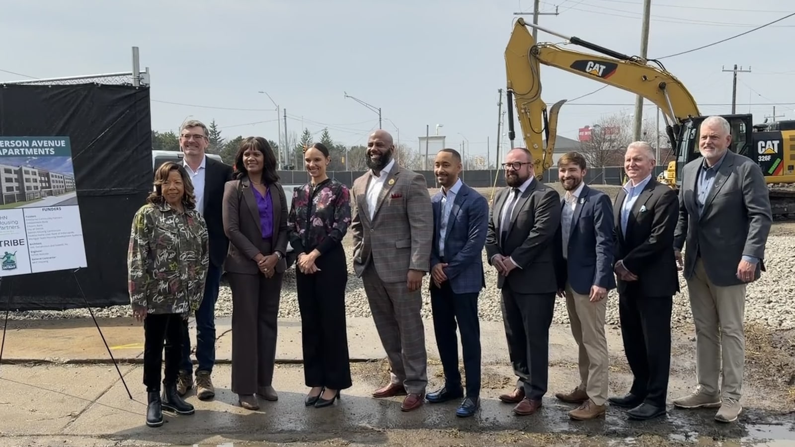 Photograph of nine people standing on a gravel construction site in front of a yellow excavator, beside an easel with a poster of the planned building